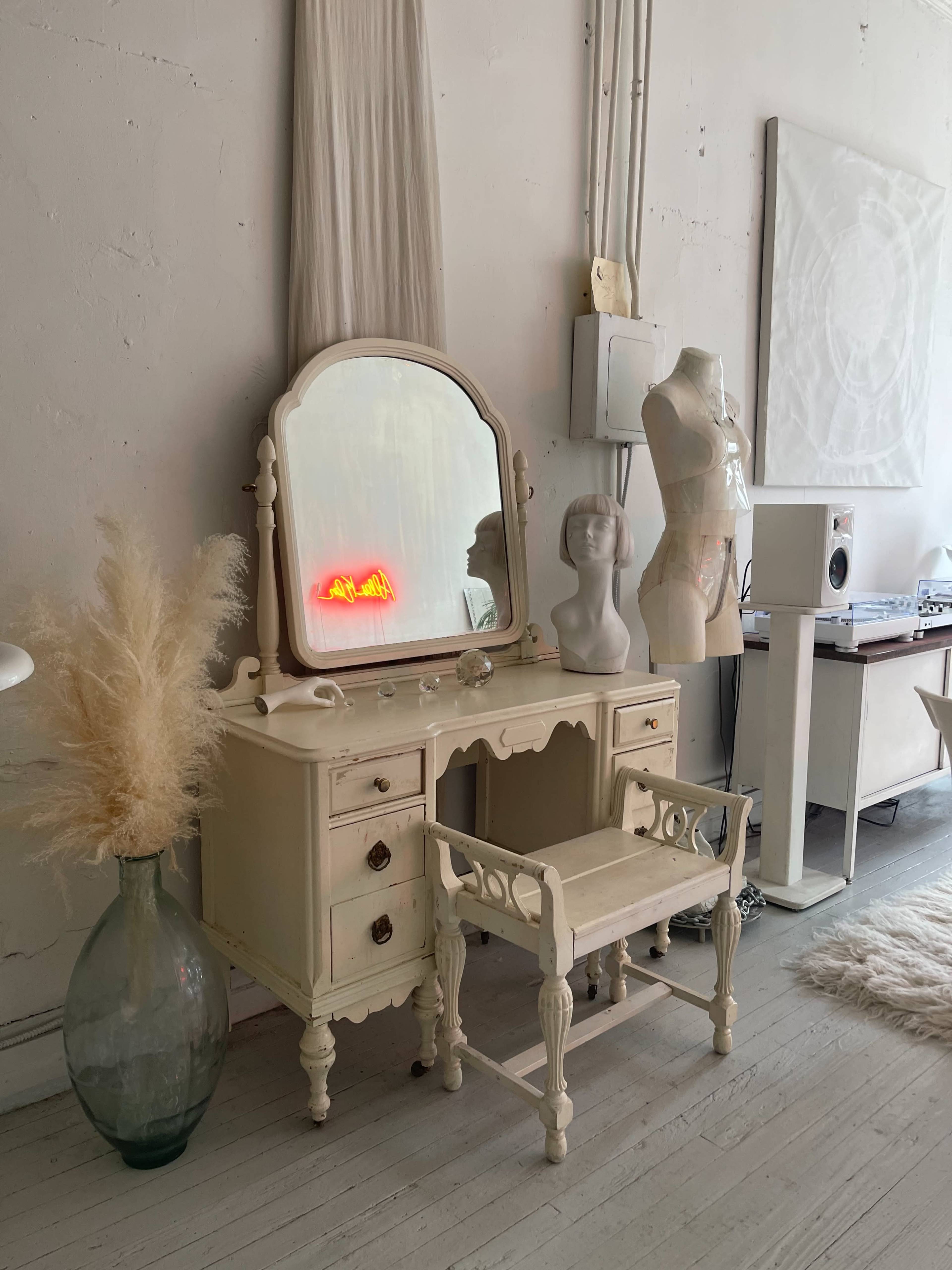 A vintage vanity table with a large mirror and a mannequin stands against a light-colored wall, accompanied by decorative items including pampas grass in a vase and audio equipment.