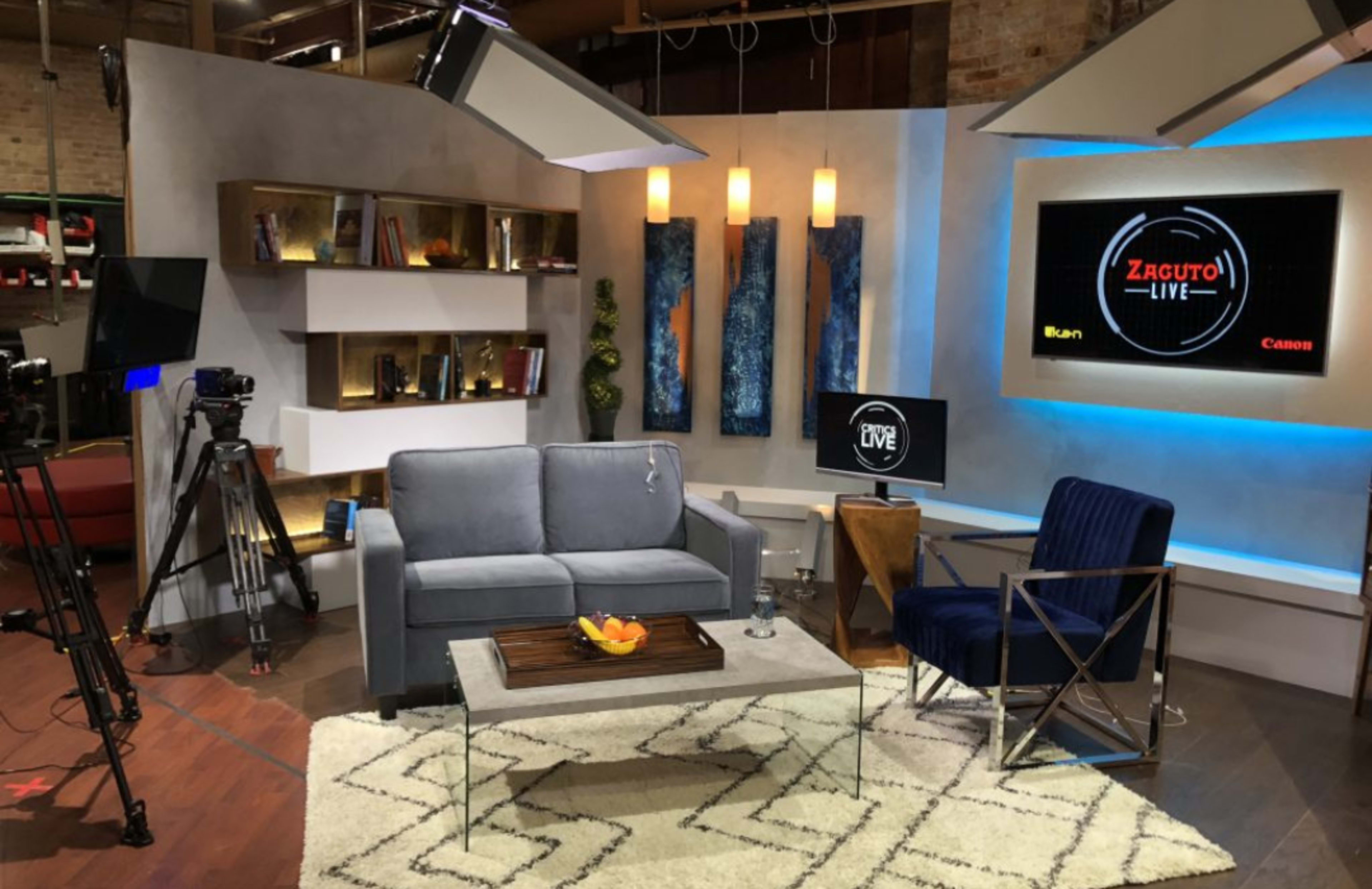 A television studio set with a gray sofa, a blue armchair, a coffee table with fruit, and multiple cameras and lights positioned around the space.