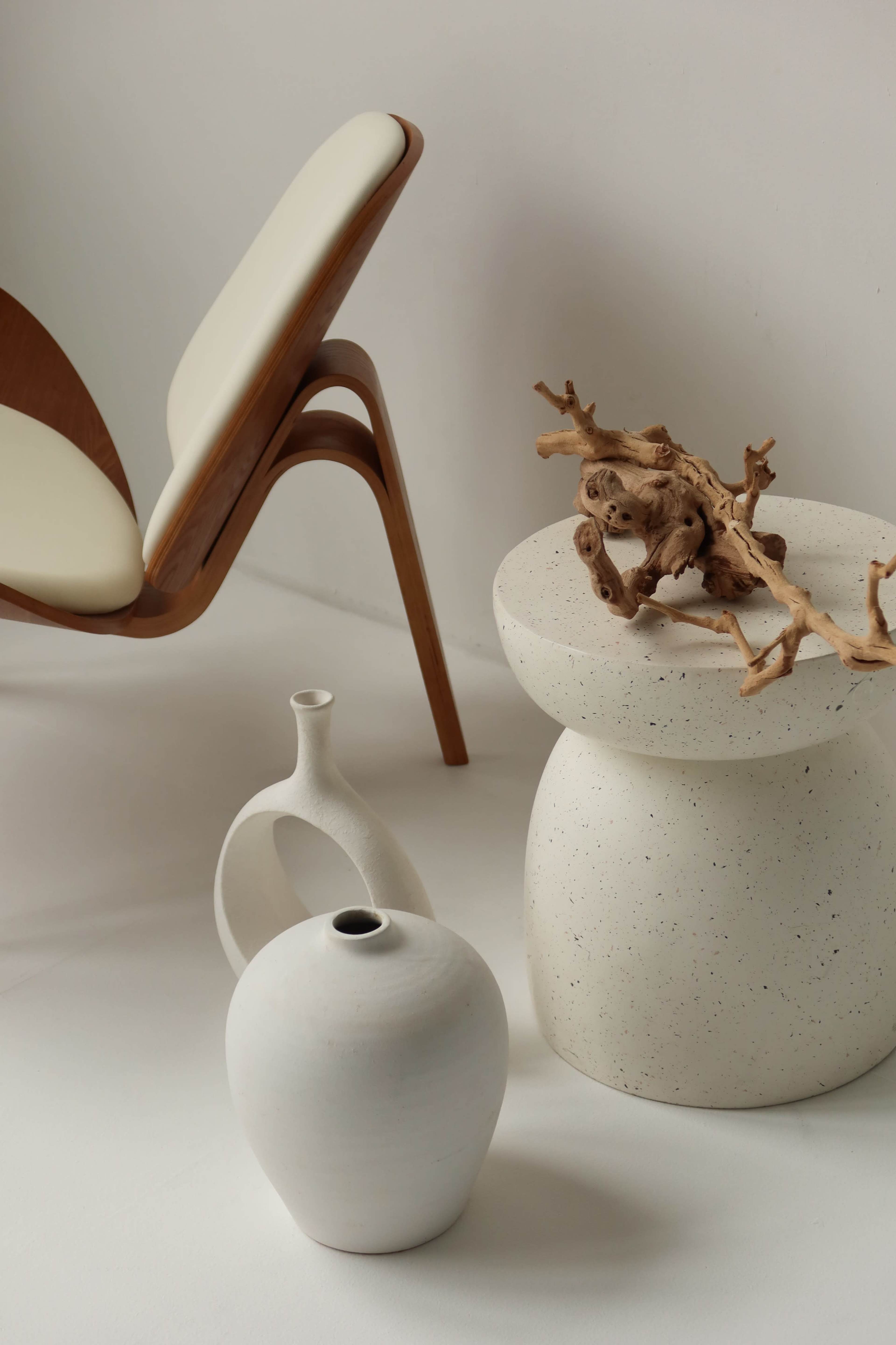 A minimalist scene features a wooden chair, a round speckled table, and two ceramic vases displayed against a neutral backdrop.