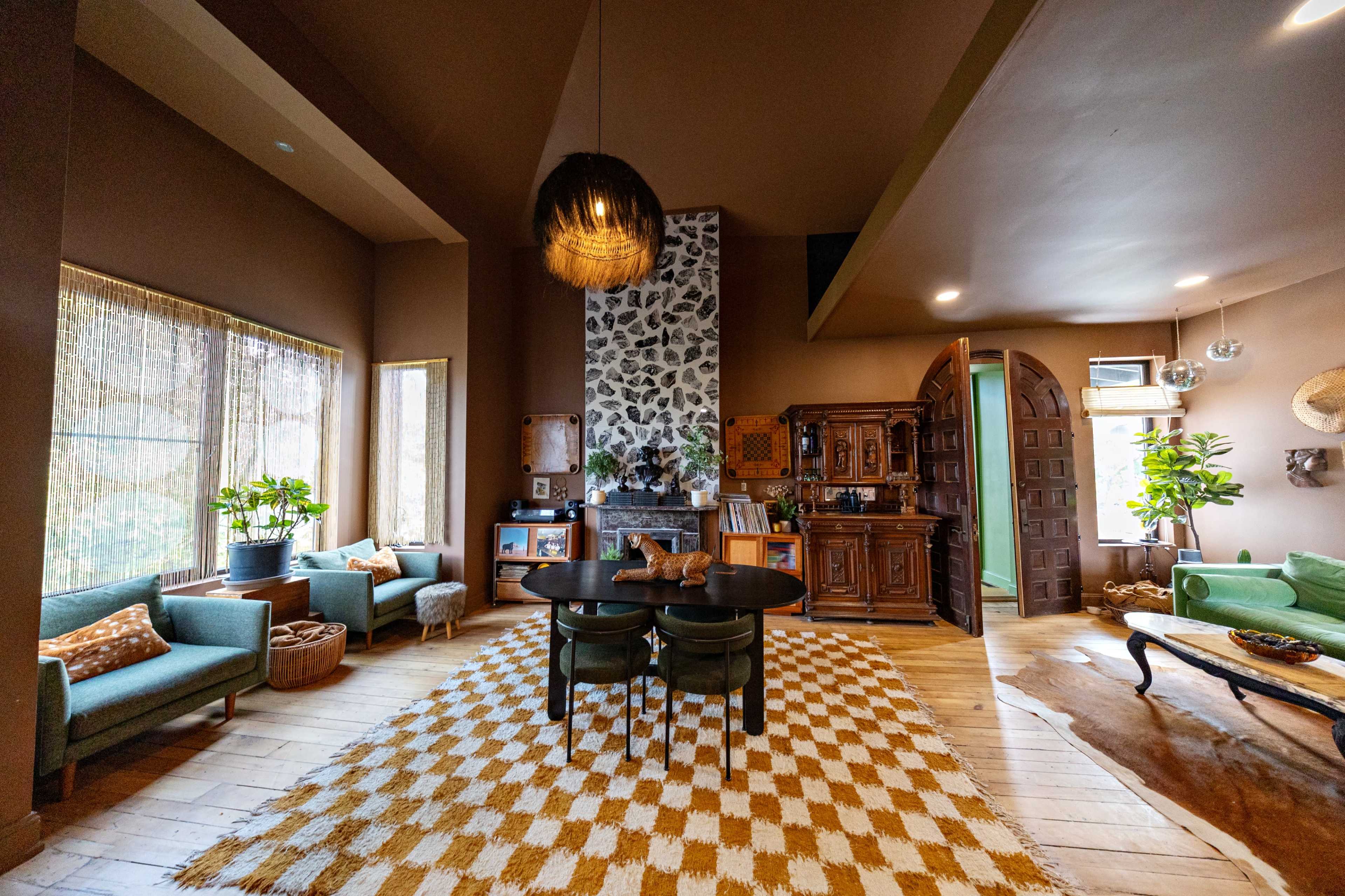 The image shows a spacious living room with a round dining table at the center, surrounded by various seating areas and a stone fireplace.