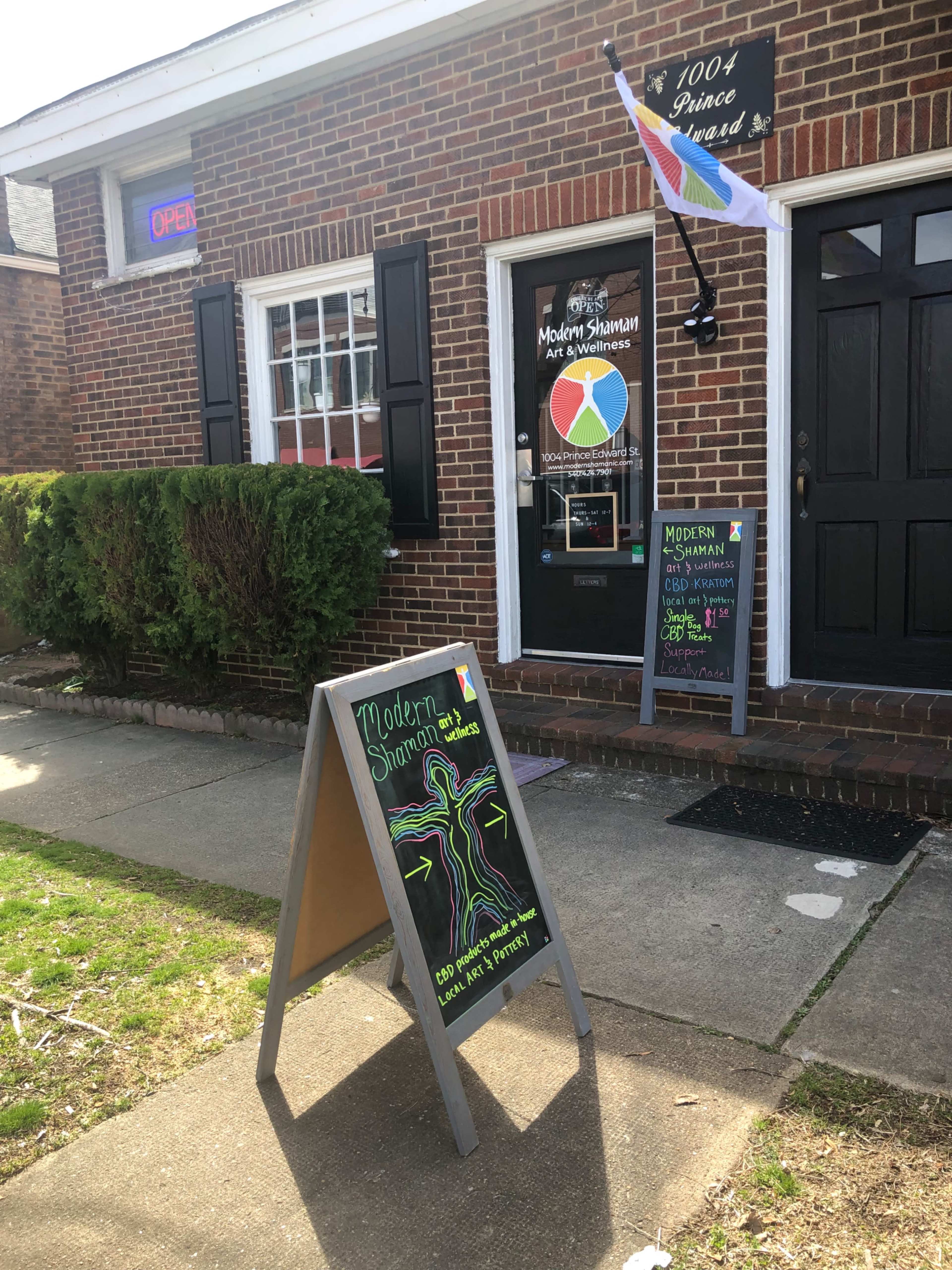 The exterior of a brick building features an open sign, an artistic storefront display, and a sandwich board with colorful chalk lettering advertising services.
