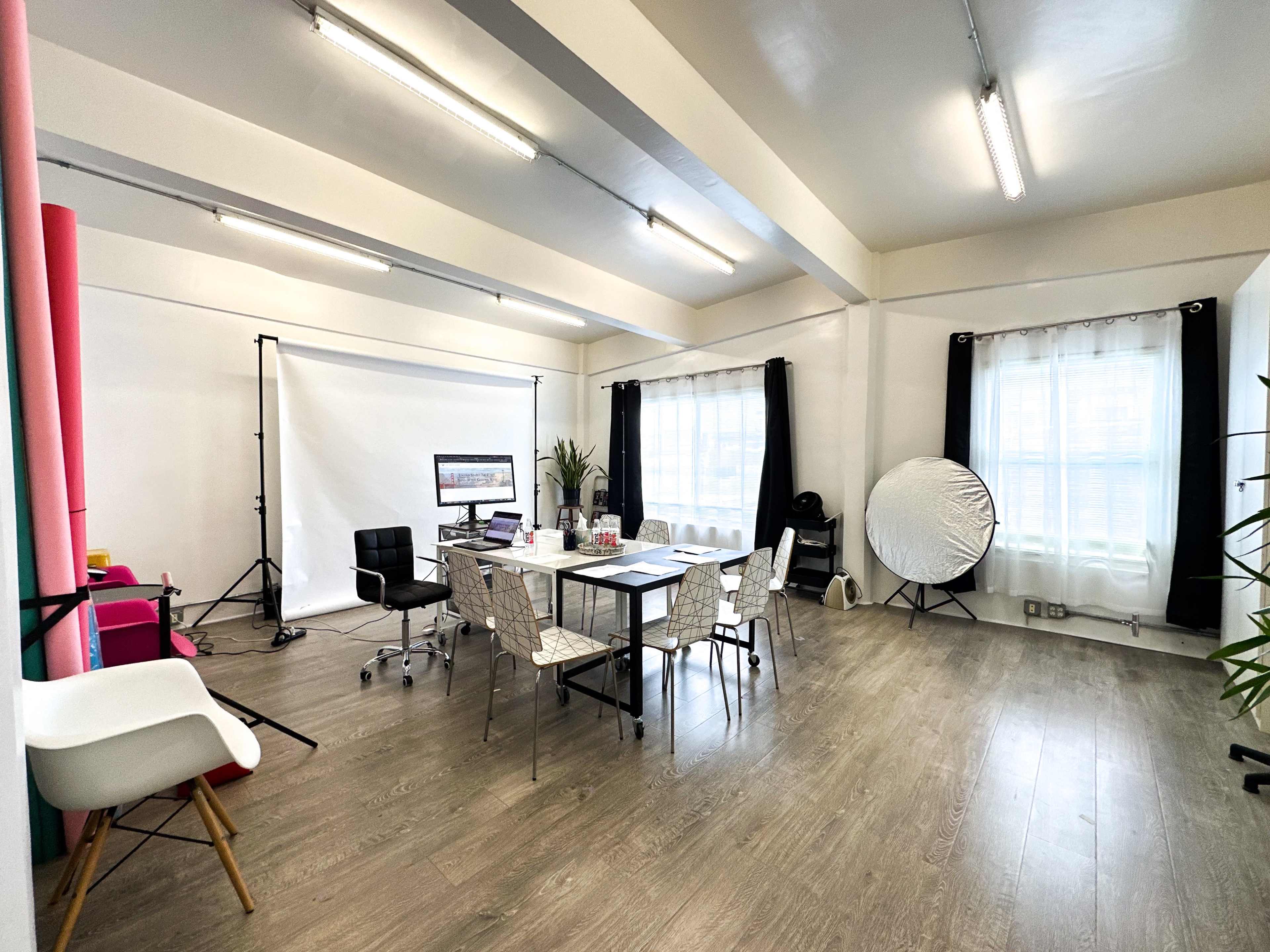 A spacious meeting room with a large table surrounded by chairs, equipped with a projector and a backdrop, and illuminated by natural light through large windows.