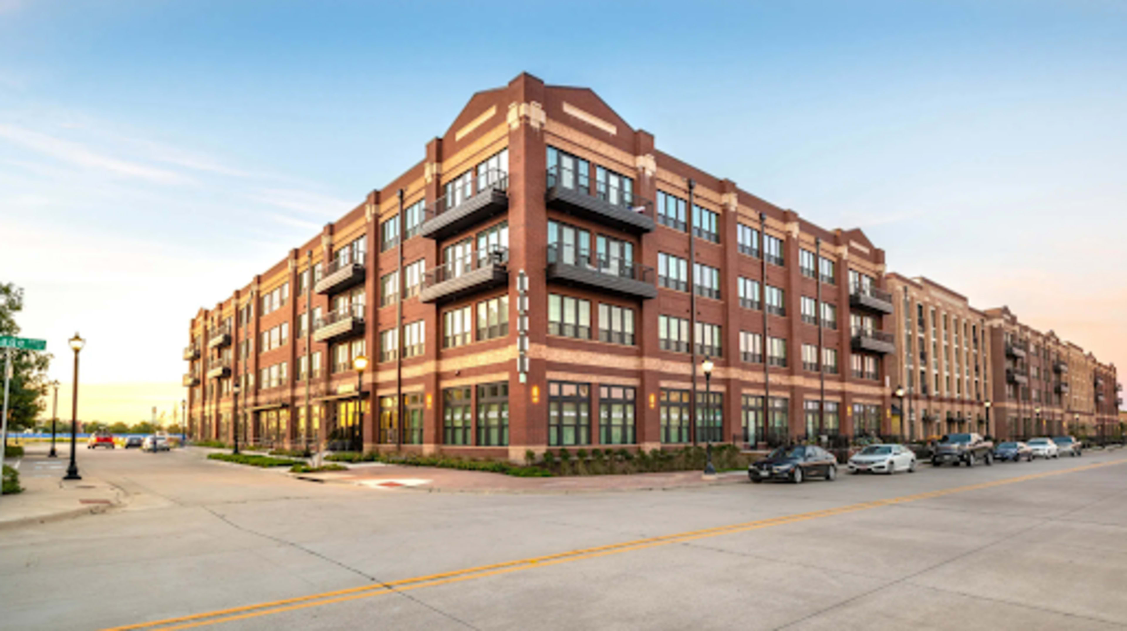 A brick apartment building with several balconies is situated at the corner of a street in a suburban area.