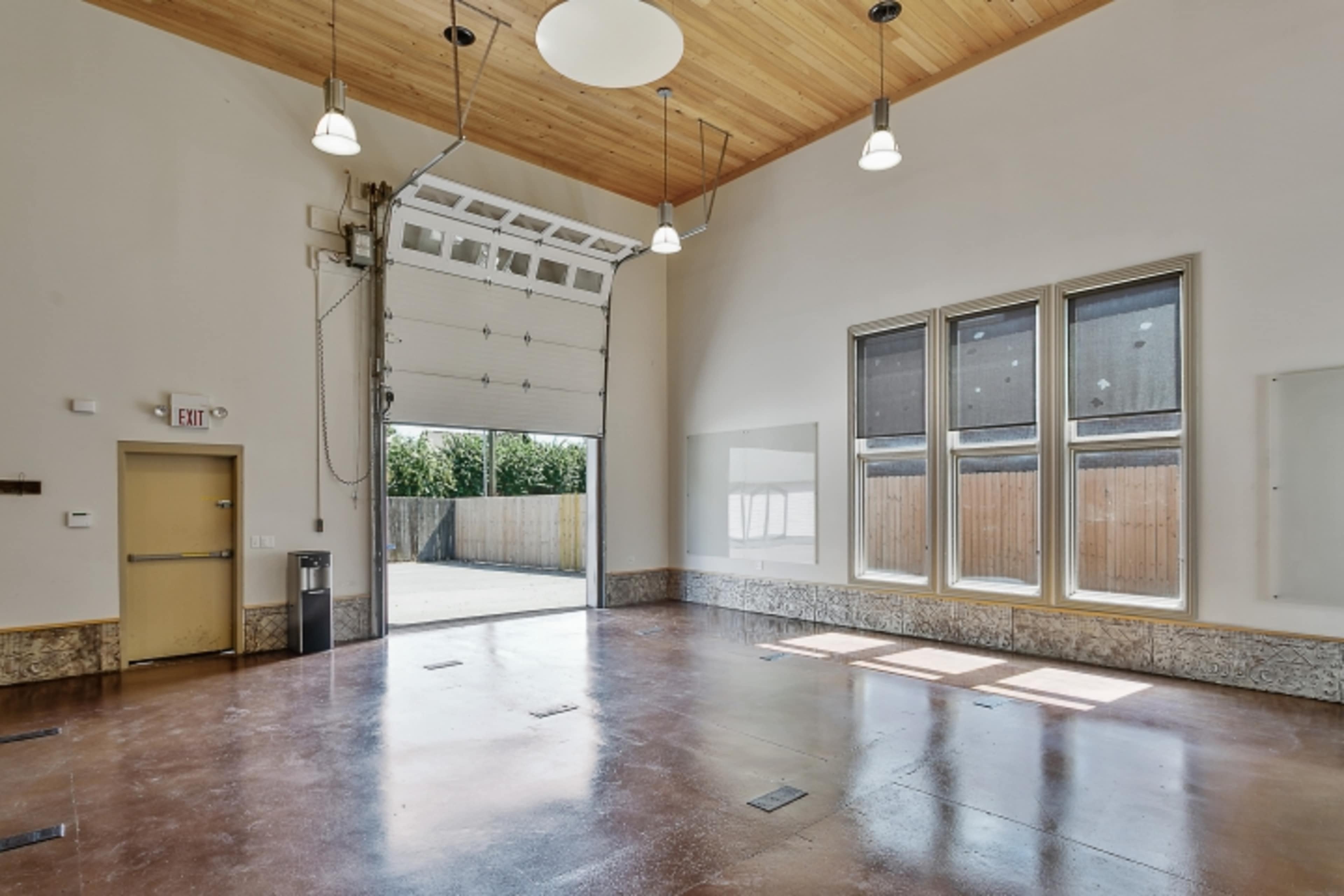 The image shows an empty interior space with a high ceiling, a large overhead door, and multiple windows letting in natural light.