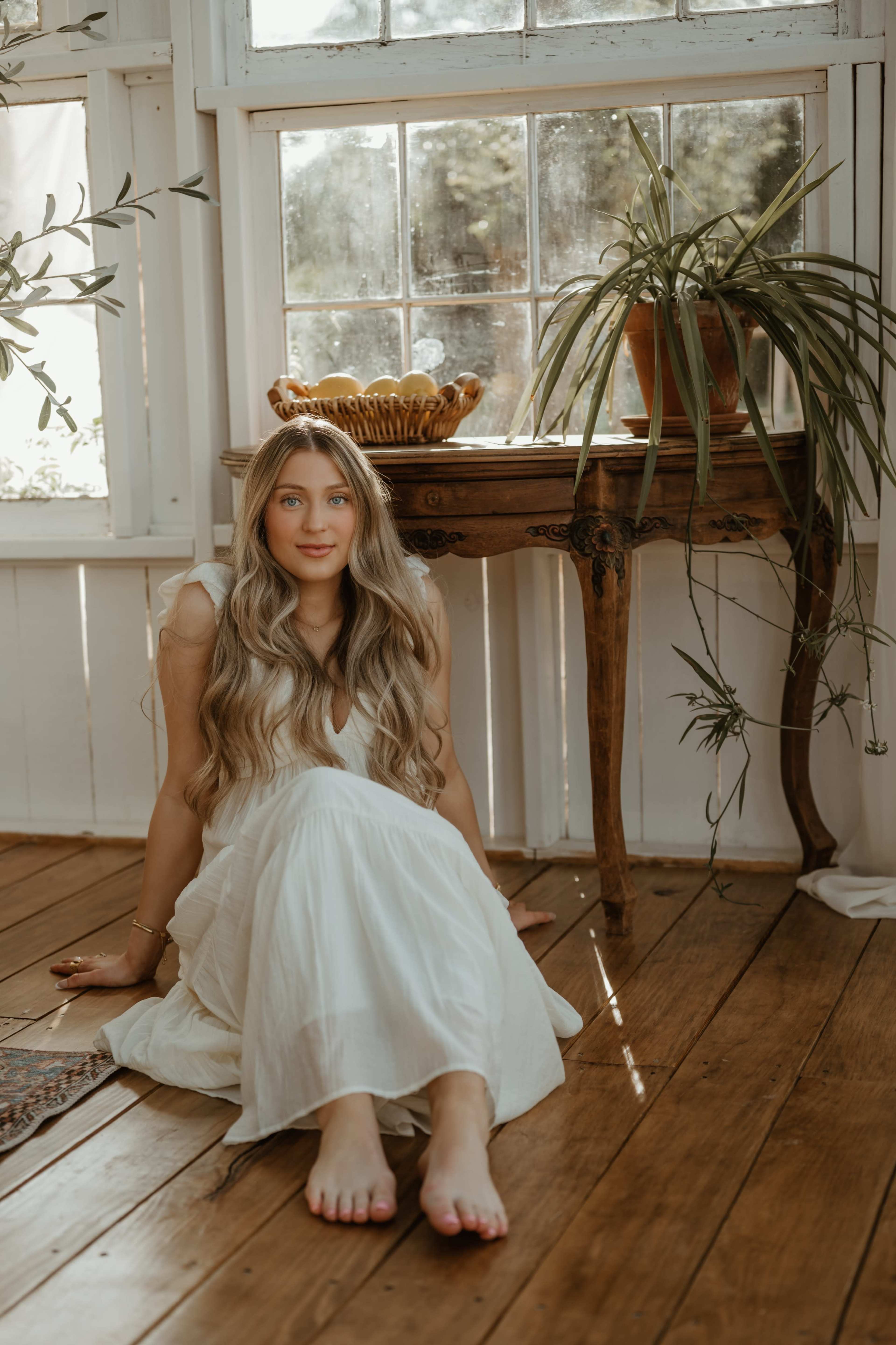 A woman sits on a wooden floor near a table with a decorative bowl, surrounded by plants and sunlight filtering through windows.