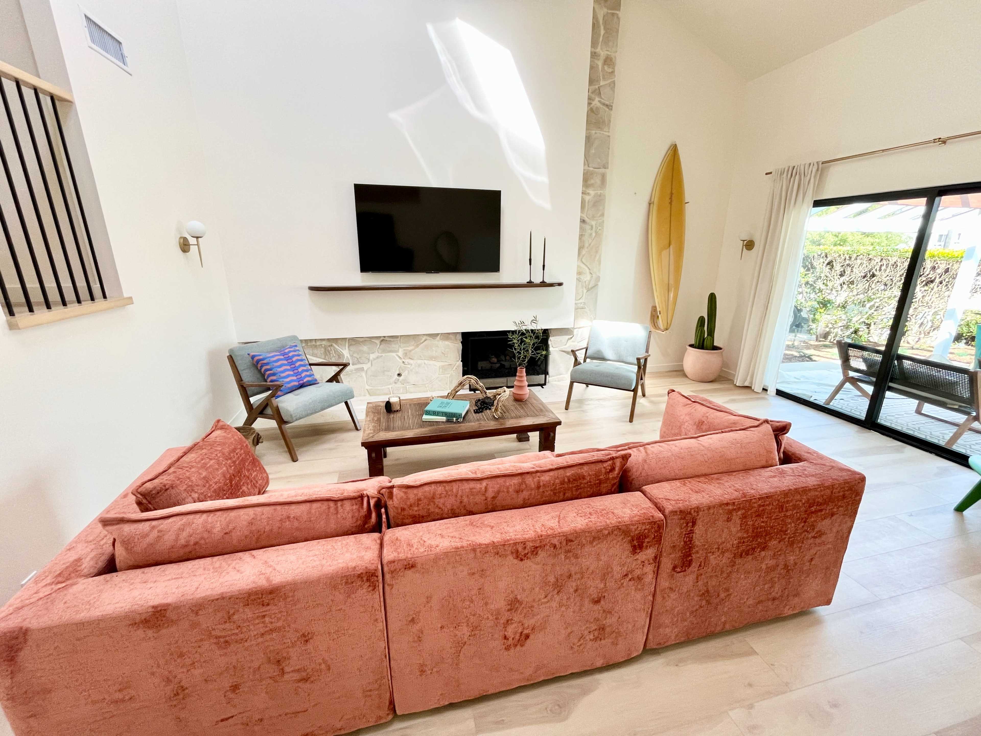 The living room features a pink sectional sofa facing a stone fireplace with a wall-mounted TV, surrounded by two chairs and a view of a patio with plants.