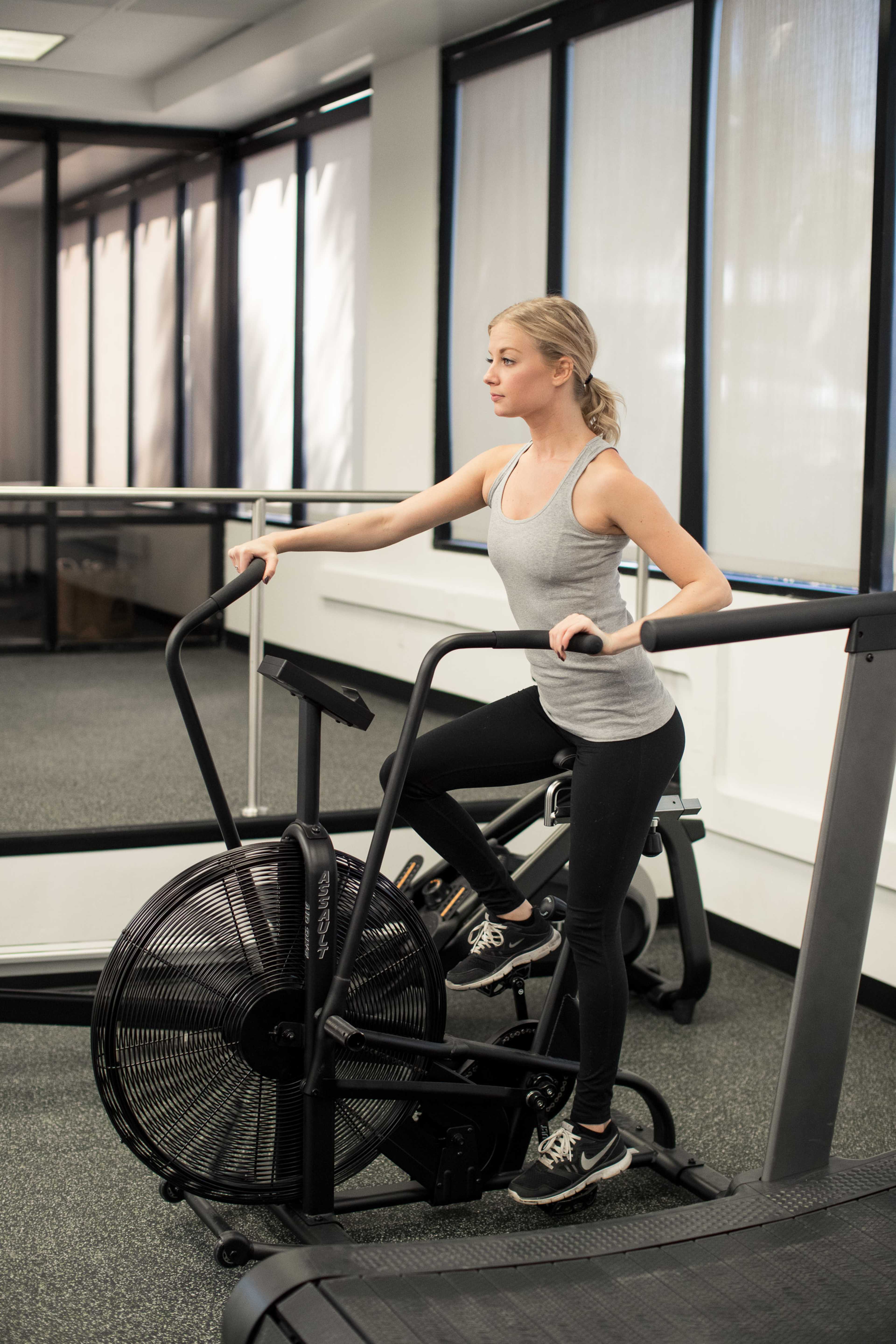 A young woman exercises on a stationary bike in a gym with large windows.