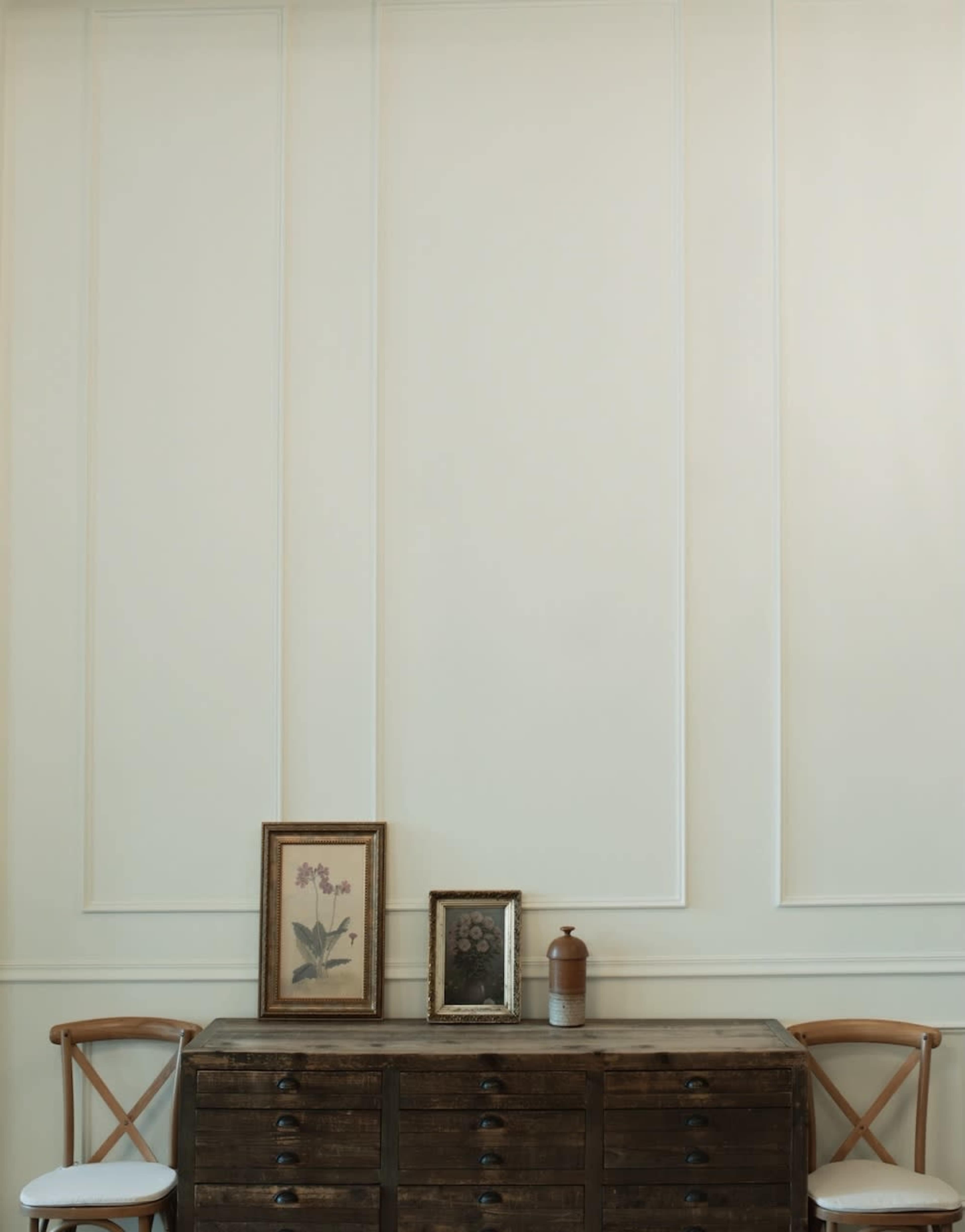 A wooden dresser is positioned against a plain, light-colored wall, featuring two framed pictures and a ceramic bottle displayed on top.