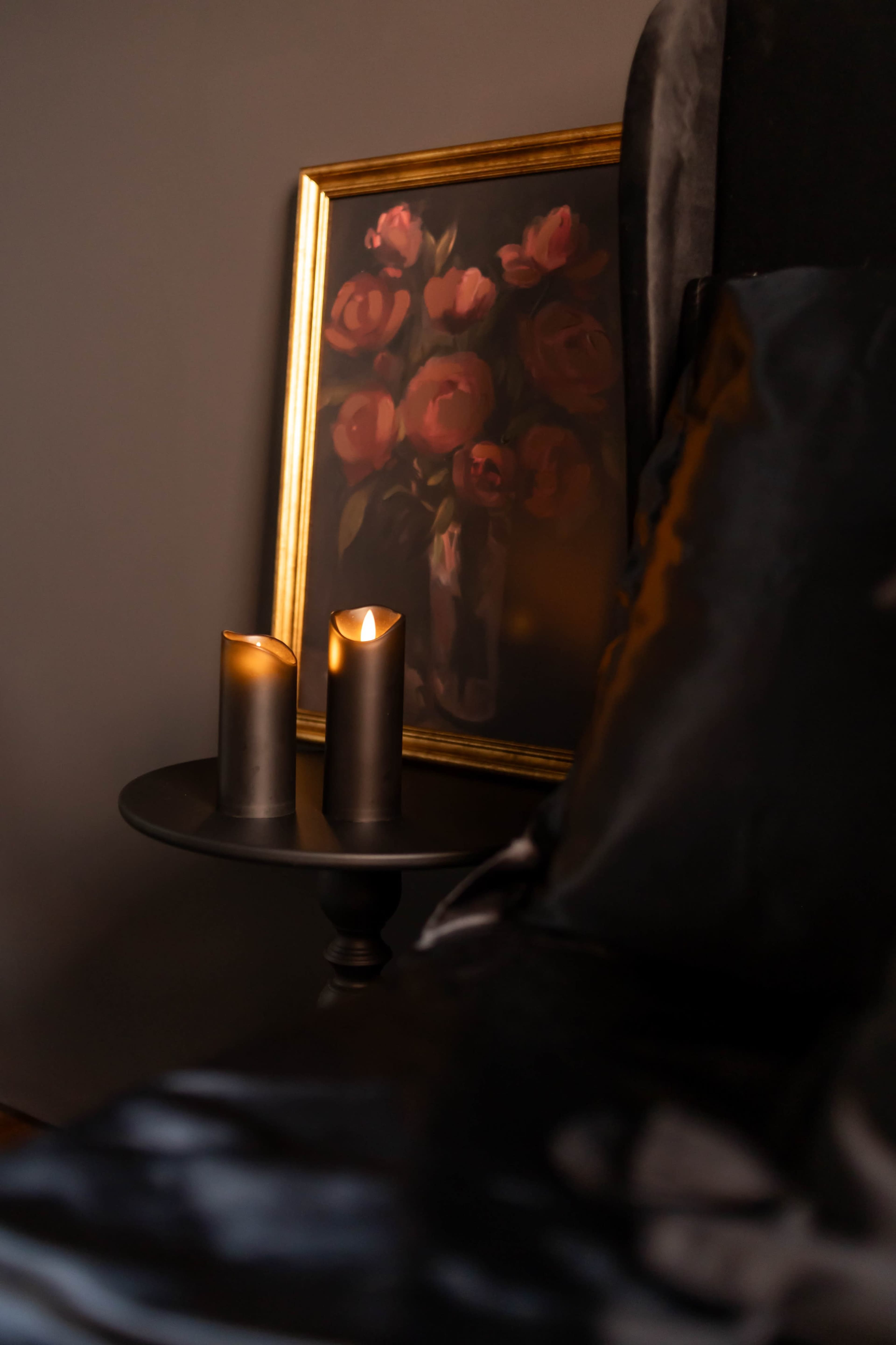 A dimly lit table with two flickering candles stands beside a framed painting of roses on a wall.