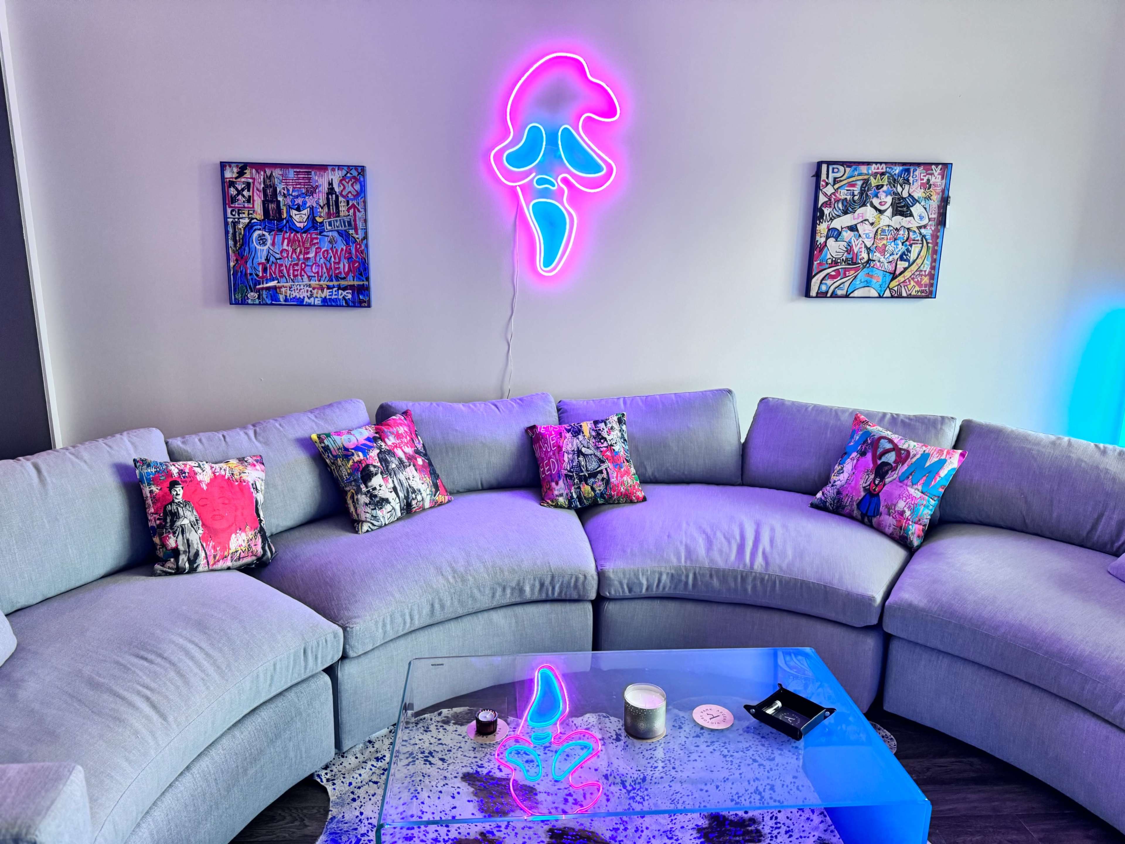 A gray sectional couch with colorful patterned pillows is positioned in a room featuring a glowing neon art piece and framed artworks on the wall.