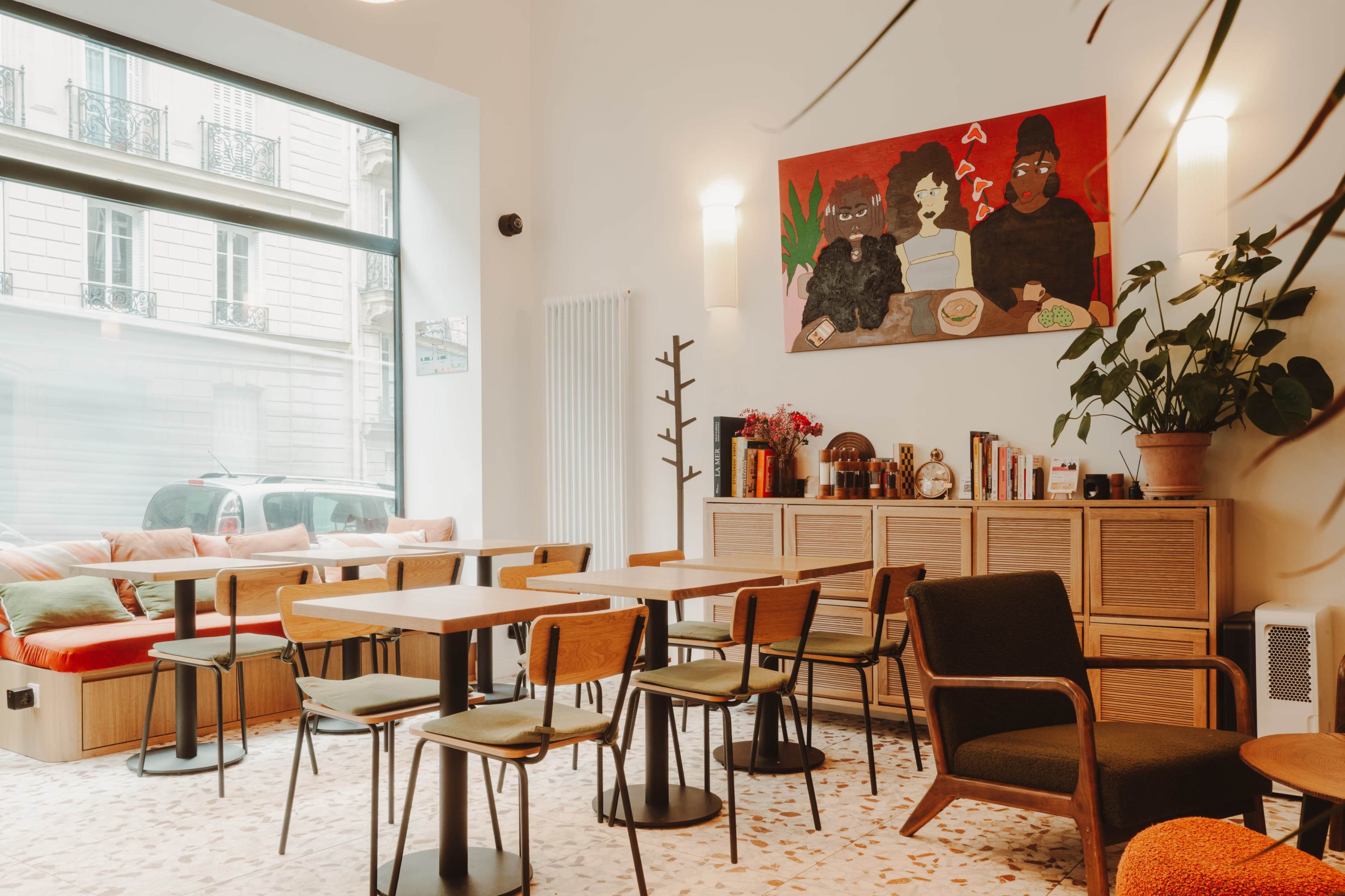 The image shows a well-lit café interior with wooden tables and chairs, a large window, and an artwork featuring three figures on the wall.