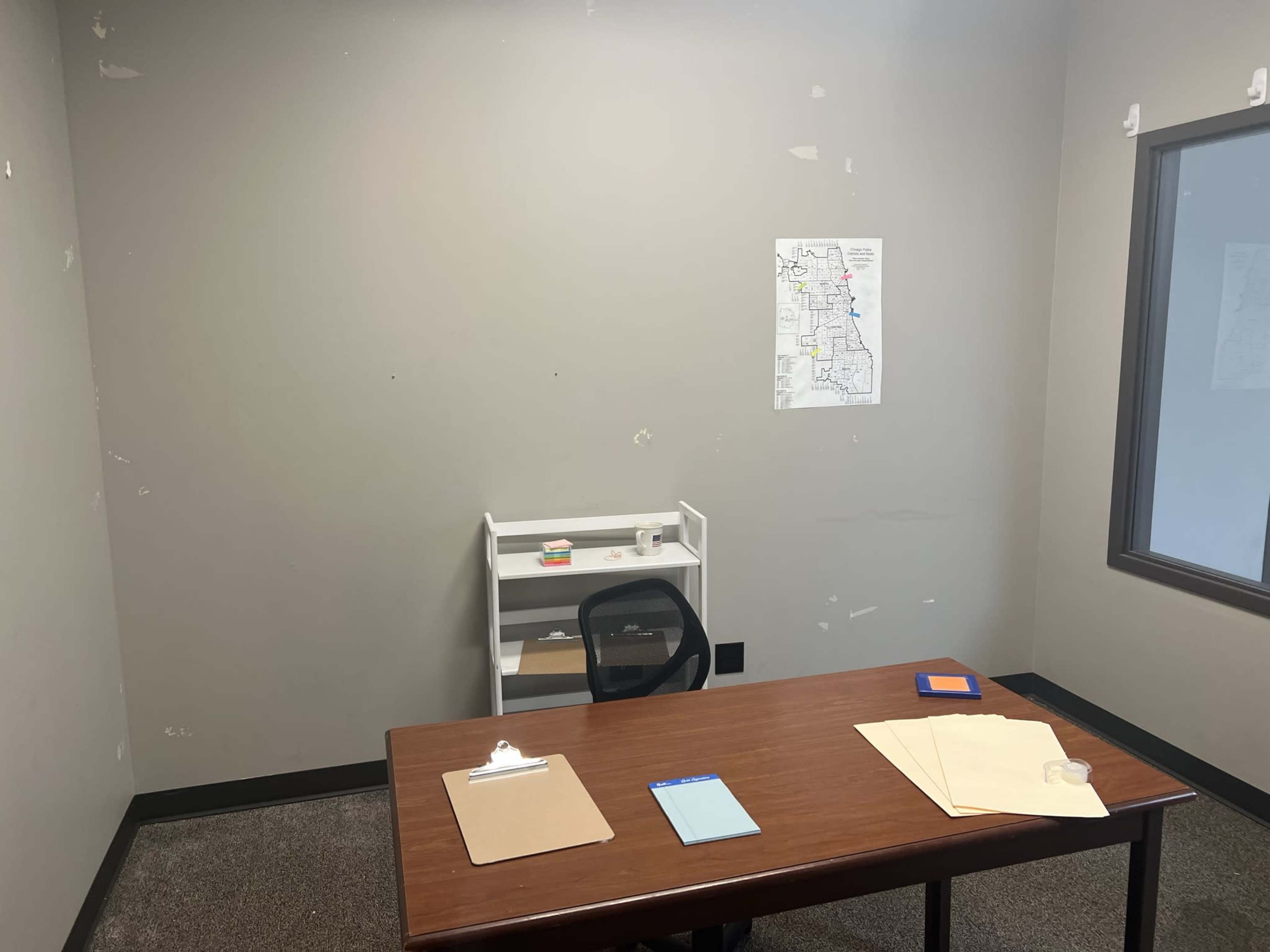 A sparsely furnished office with a wooden desk, black chair, and a wall-mounted map.