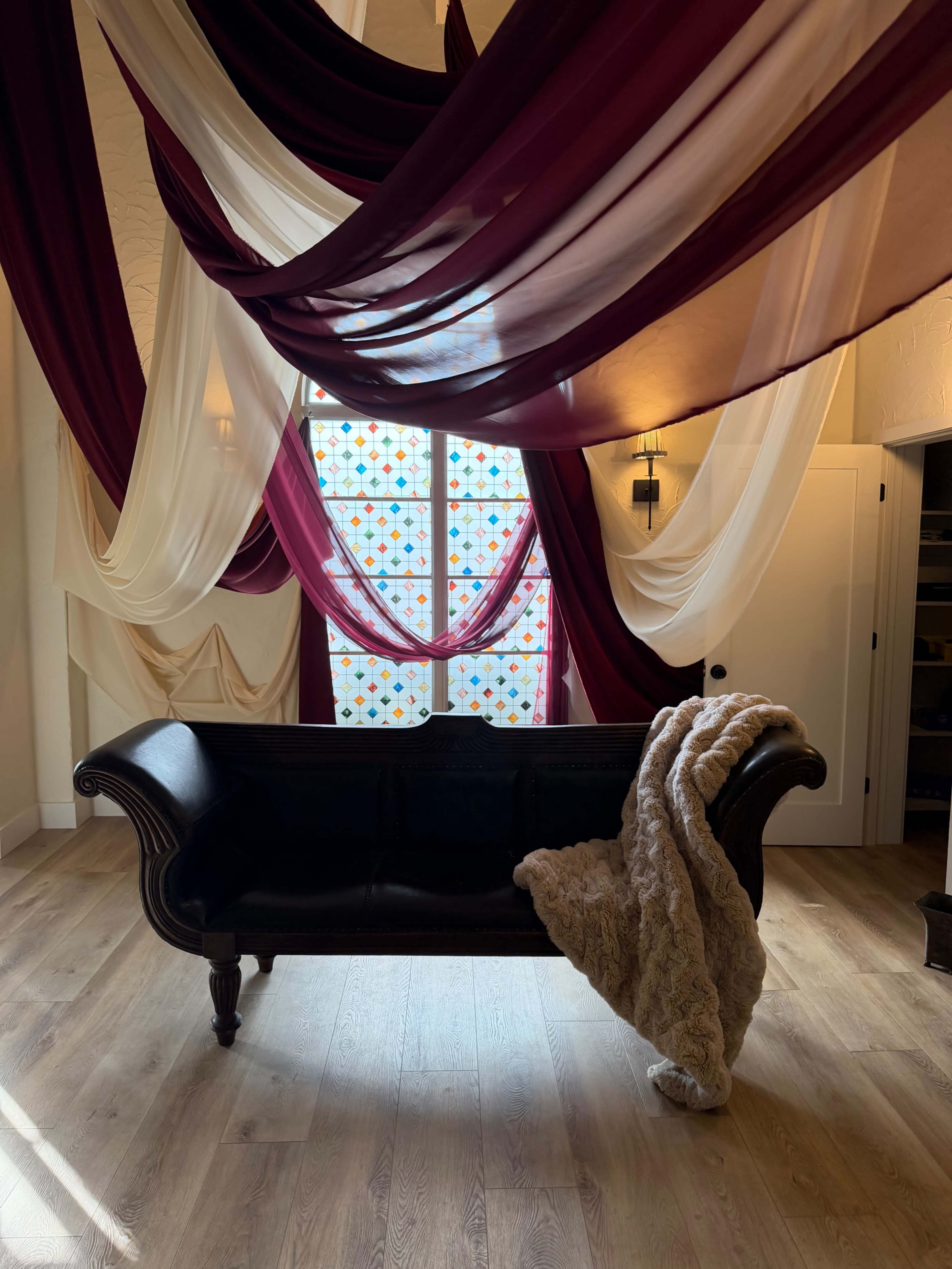 A dark couch with a soft throw is positioned in a room featuring draped fabric and a colorful stained glass window.