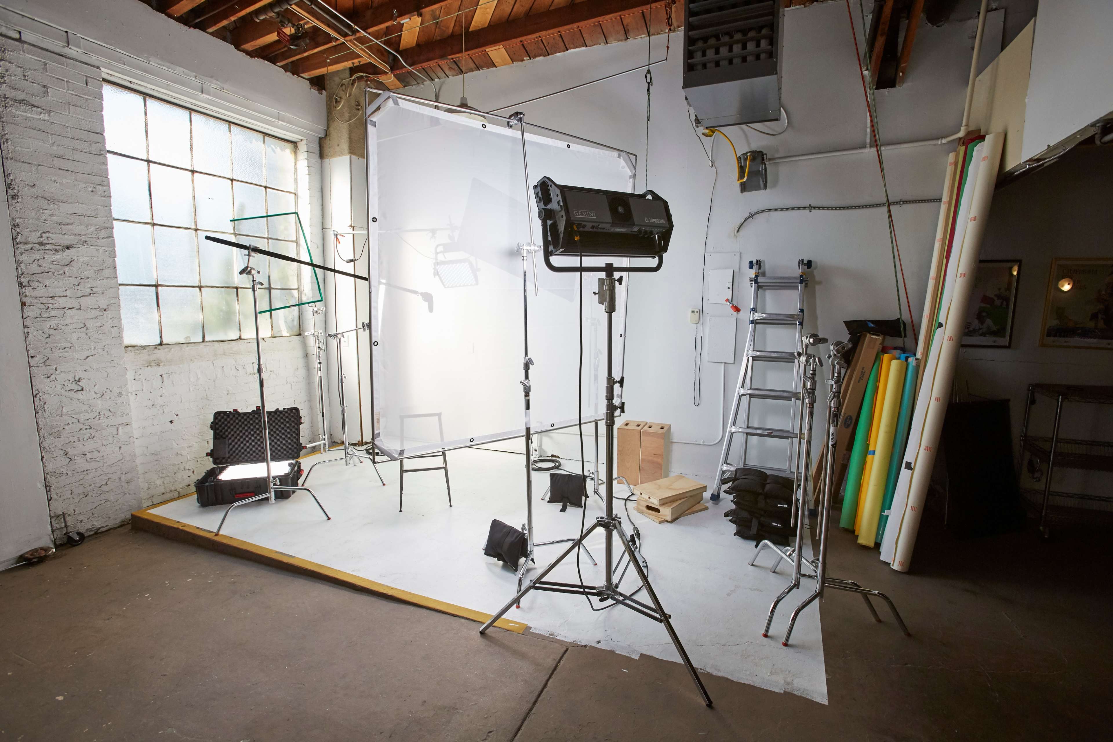 A photography studio is set up with lighting equipment, a backdrop, a ladder, and rolls of colored paper stacked against the wall.