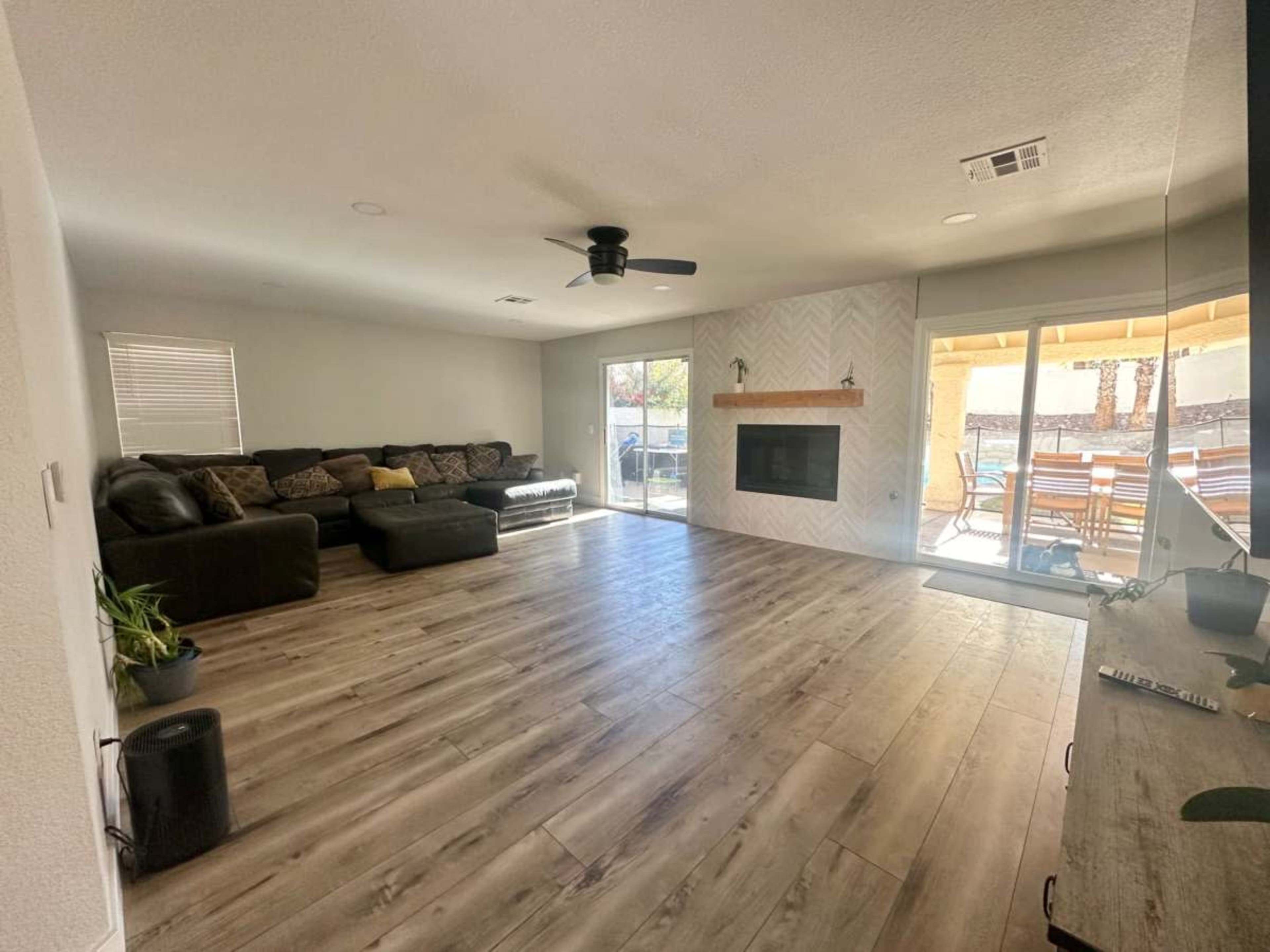 The image shows a spacious living room with a large sectional sofa, a fireplace on one wall, sliding glass doors leading to an outdoor area, and light-colored wooden flooring.