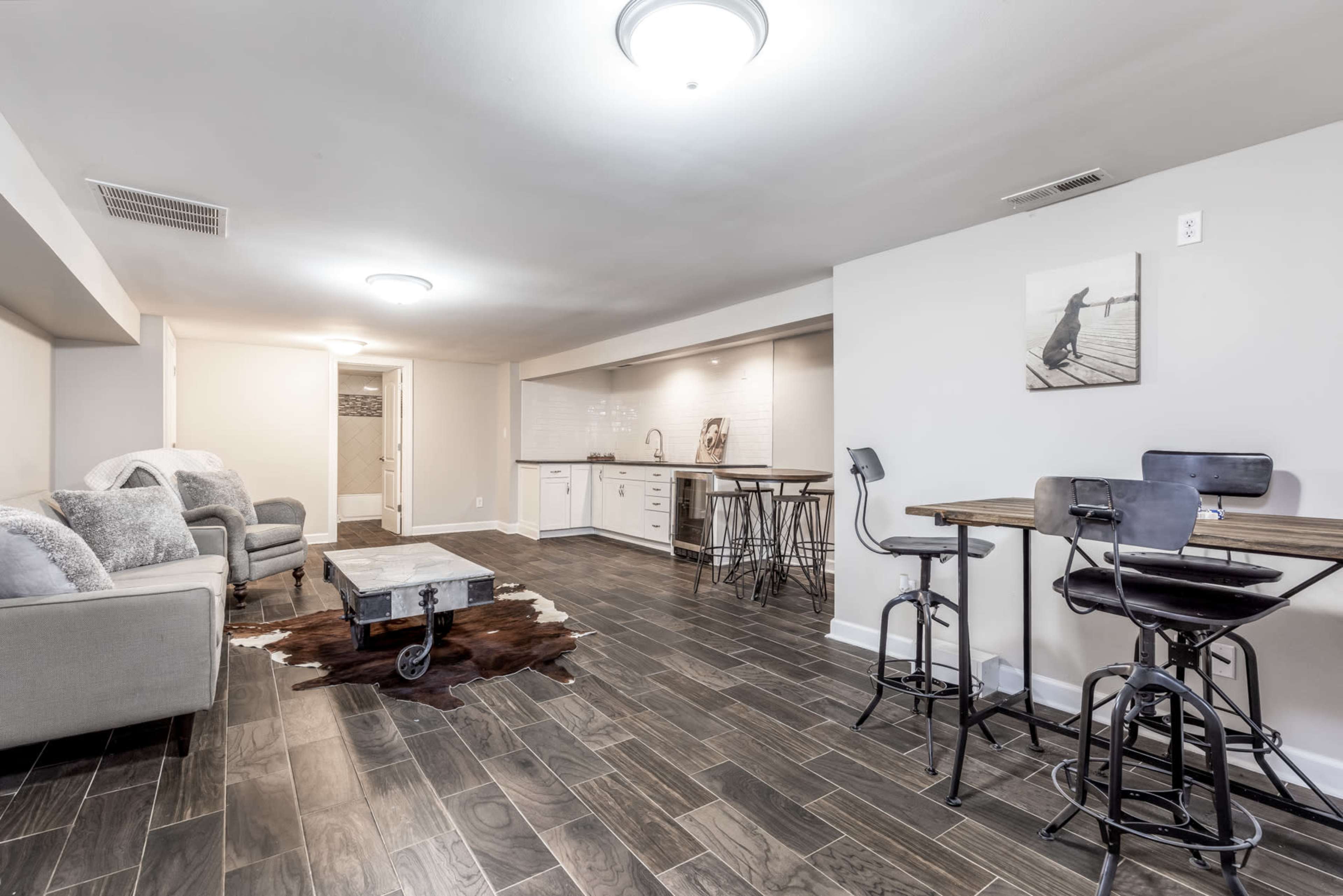 A modern basement features a seating area with a couch and two chairs, a small dining space with bar stools and a table, and a kitchenette along one wall.