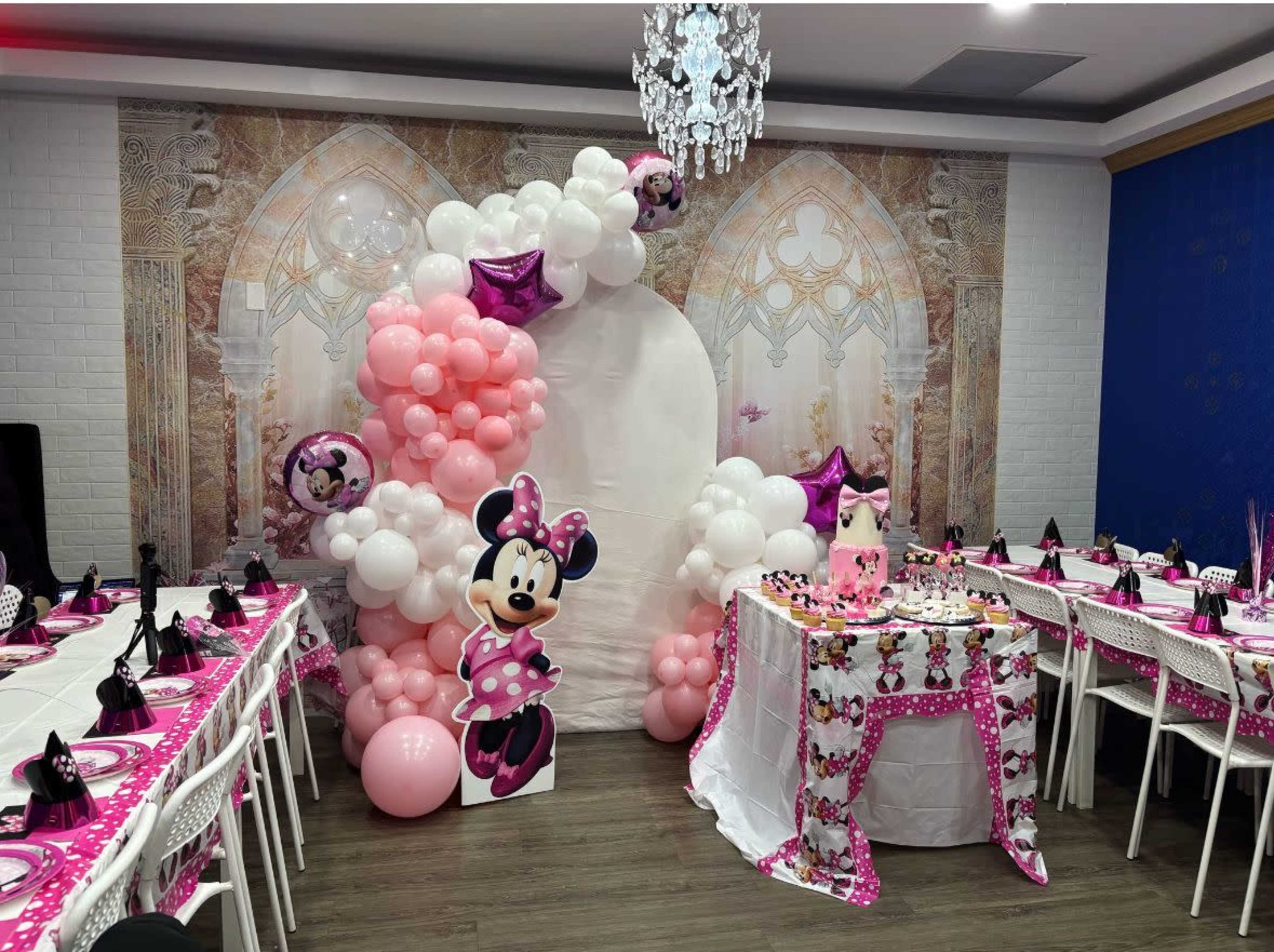 The image shows a decorated party scene featuring a large arch made of pink and white balloons, with a Minnie Mouse cutout, alongside tables set with pink tablecloths and black napkins.