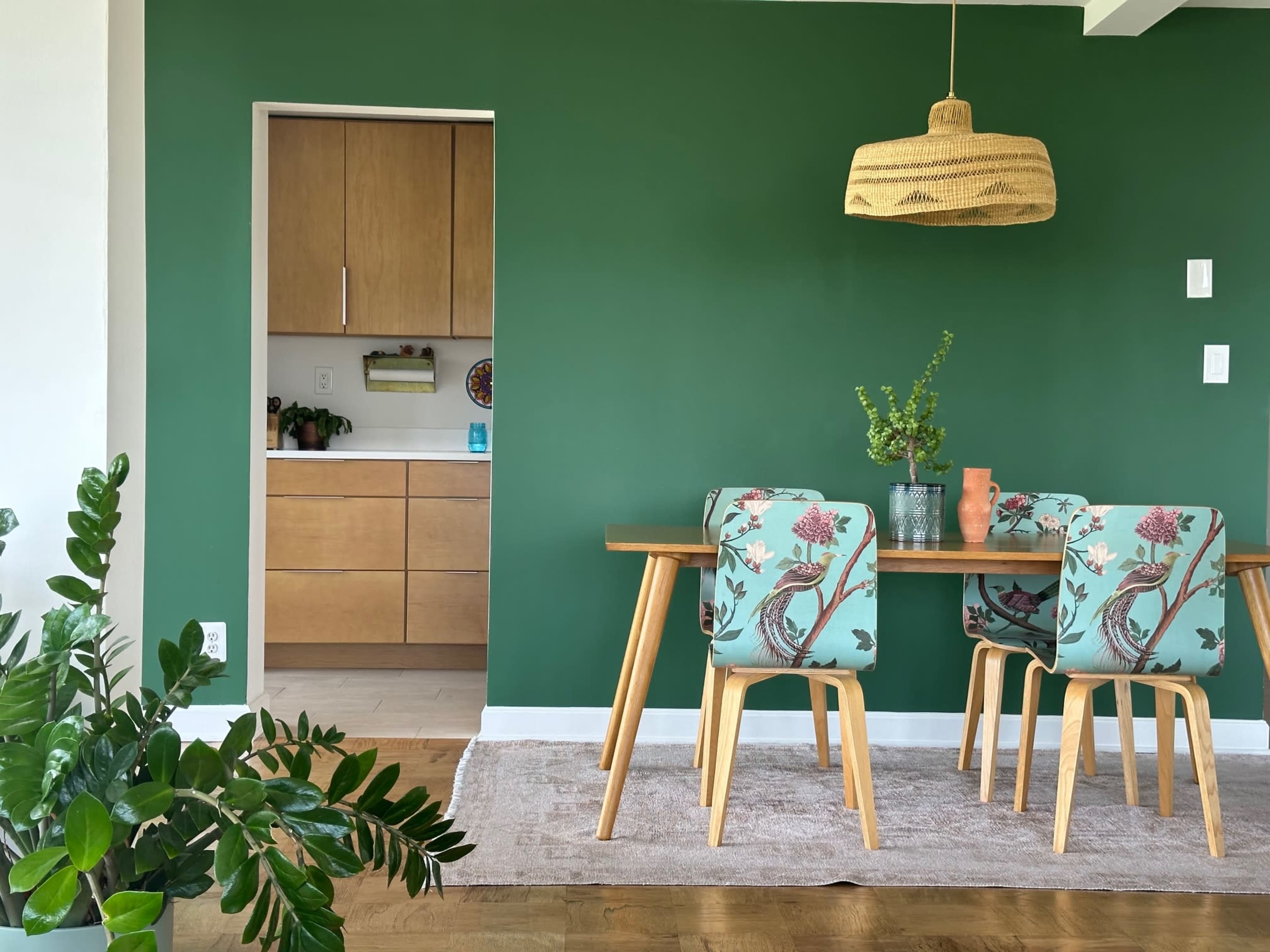 A dining area features a wooden table with patterned chairs, a green accent wall, and a woven pendant light overhead.
