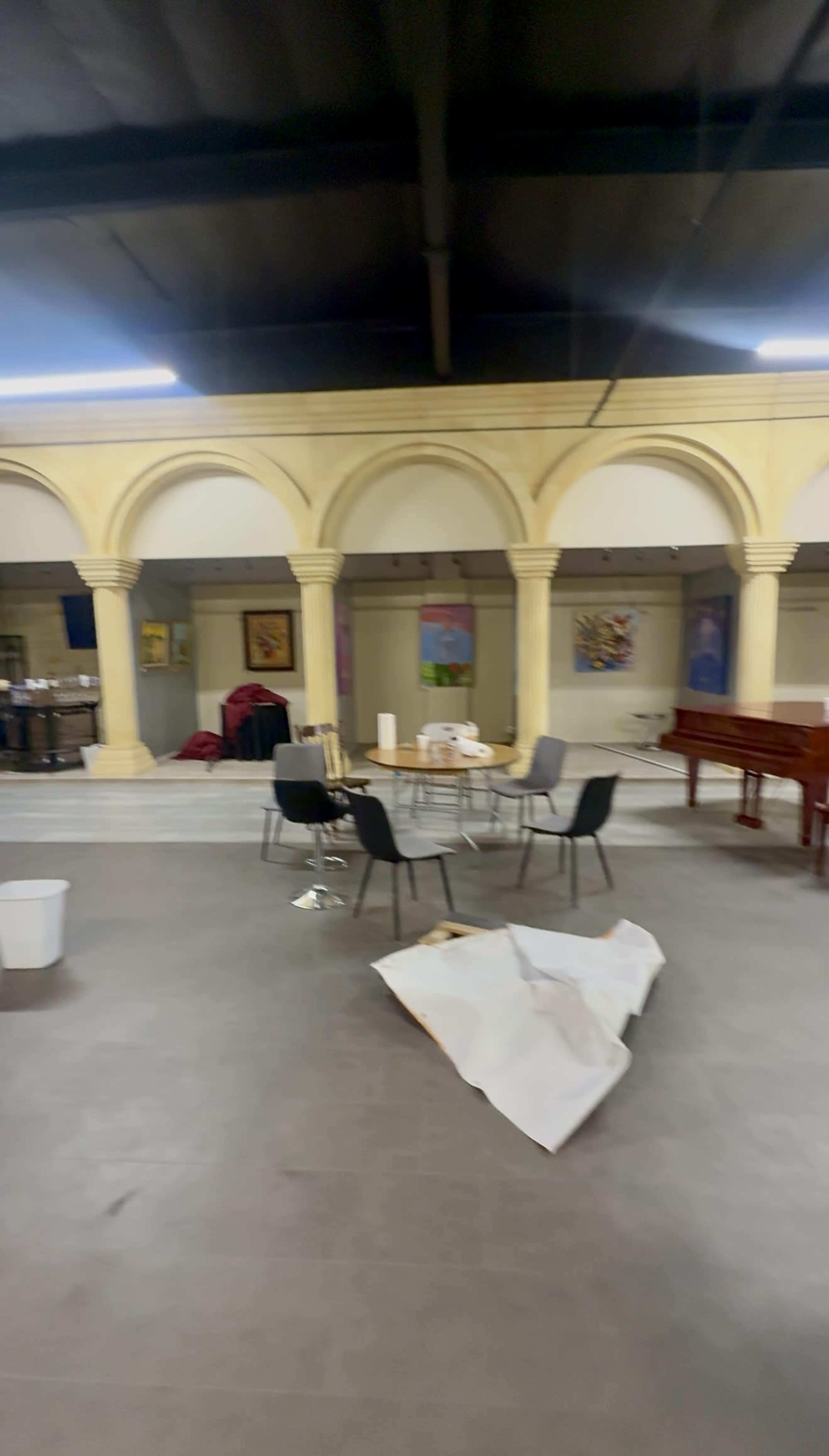The image shows a spacious indoor area with arched columns, a round table surrounded by chairs, and a piano, along with various artworks on the walls and some clutter on the floor.