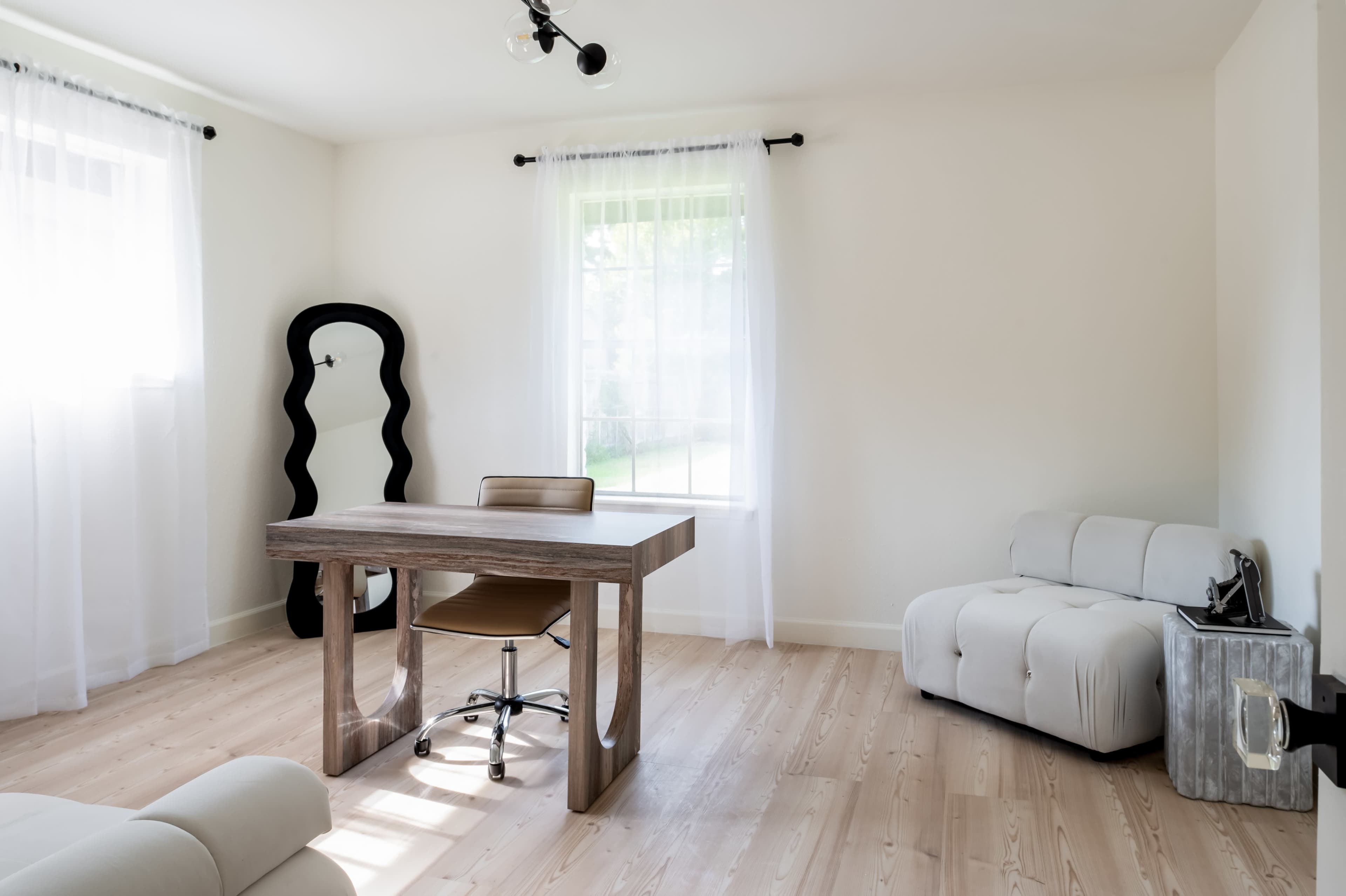 The image shows a minimalist room featuring a wooden desk with a chair, a soft couch, and large windows with sheer curtains.