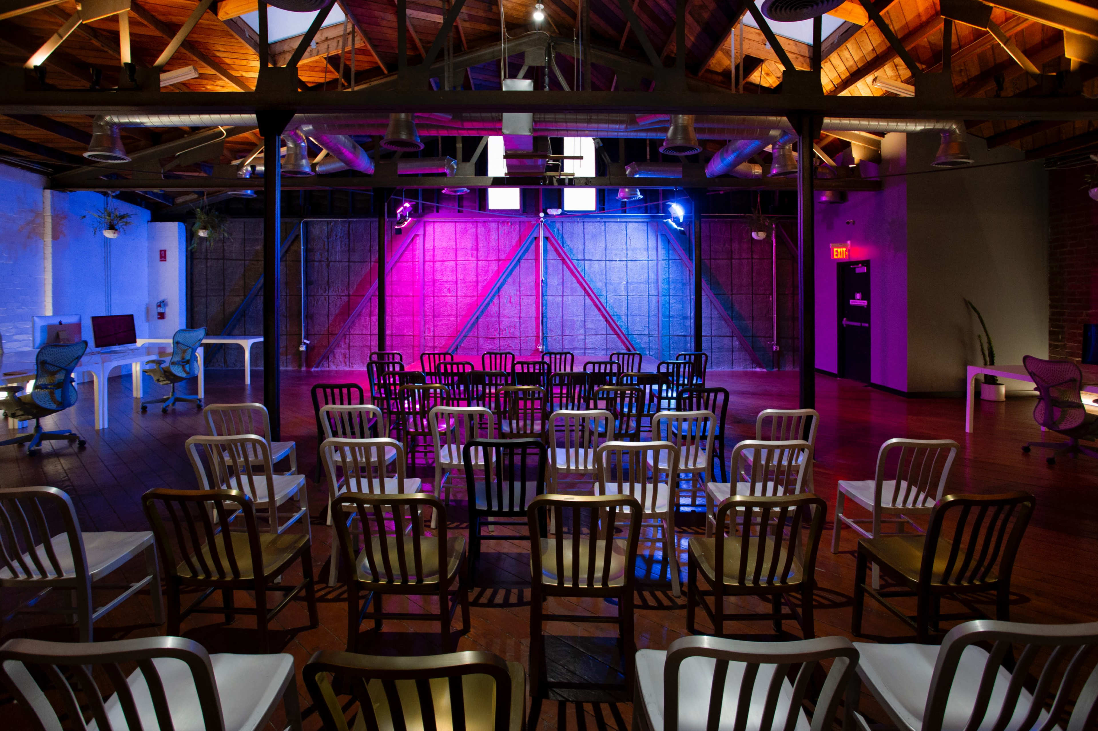 A spacious room with wooden floors and exposed beams features rows of black and white chairs facing a colorful illuminated wall.