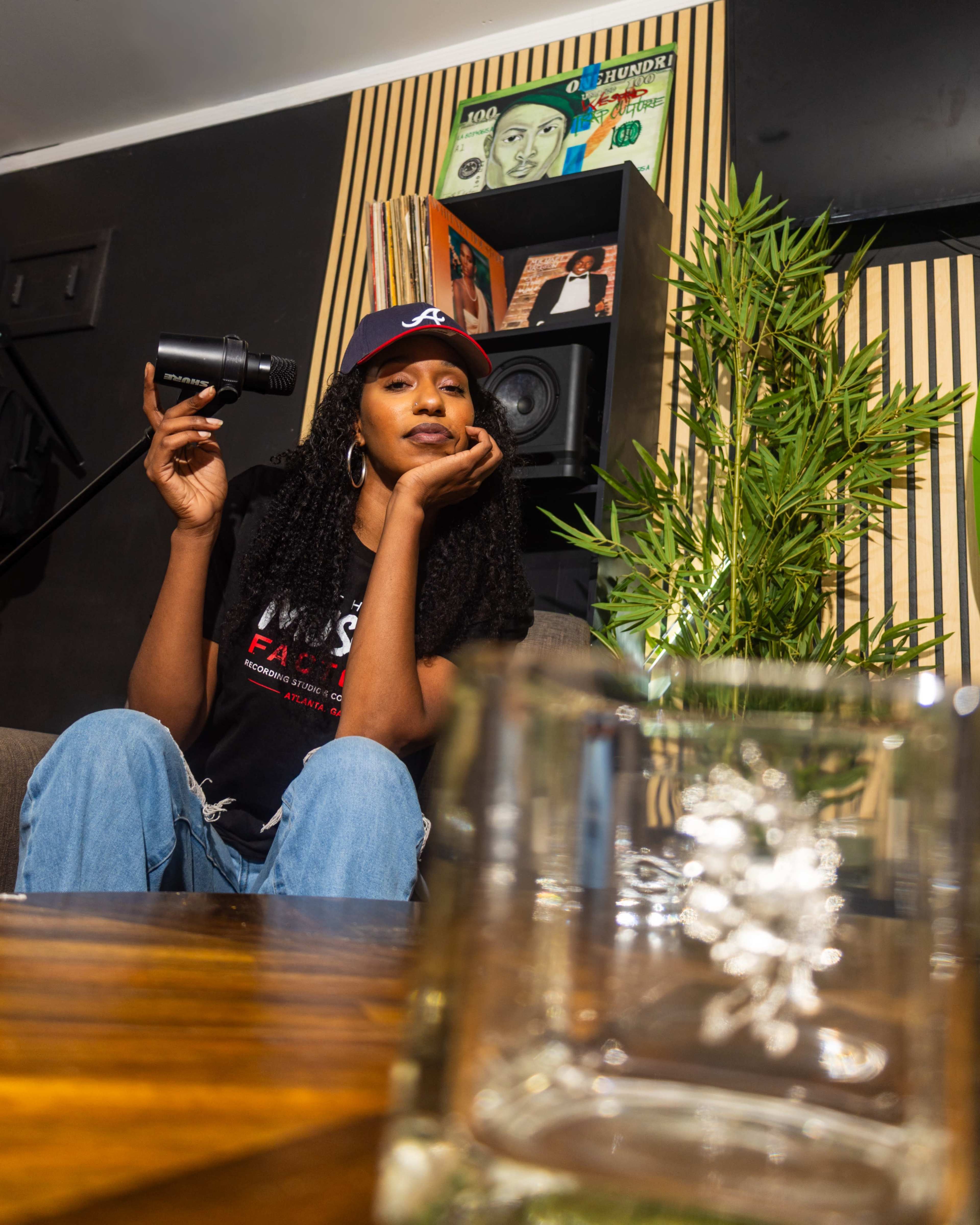 A woman sits casually on a couch with a microphone in her hand, surrounded by vinyl records and a decorative plant.