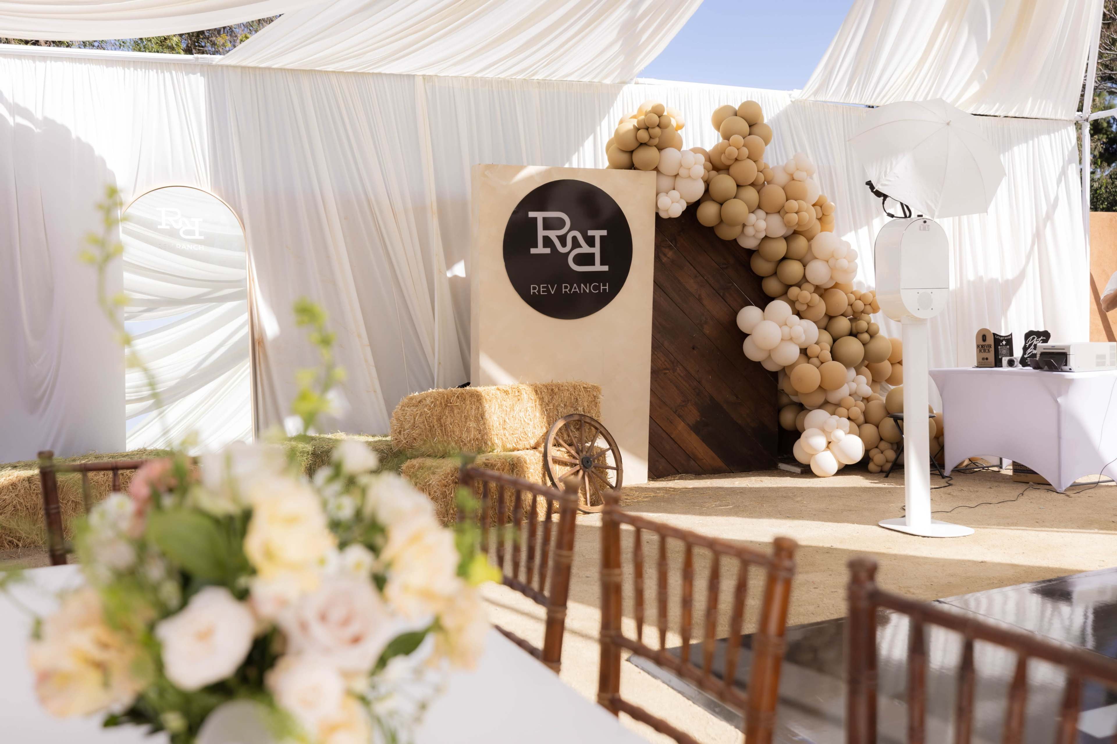 The image depicts a decorated event space featuring a backdrop with balloons, a sign for "Rev Ranch," and wooden accents, alongside a table adorned with flowers.