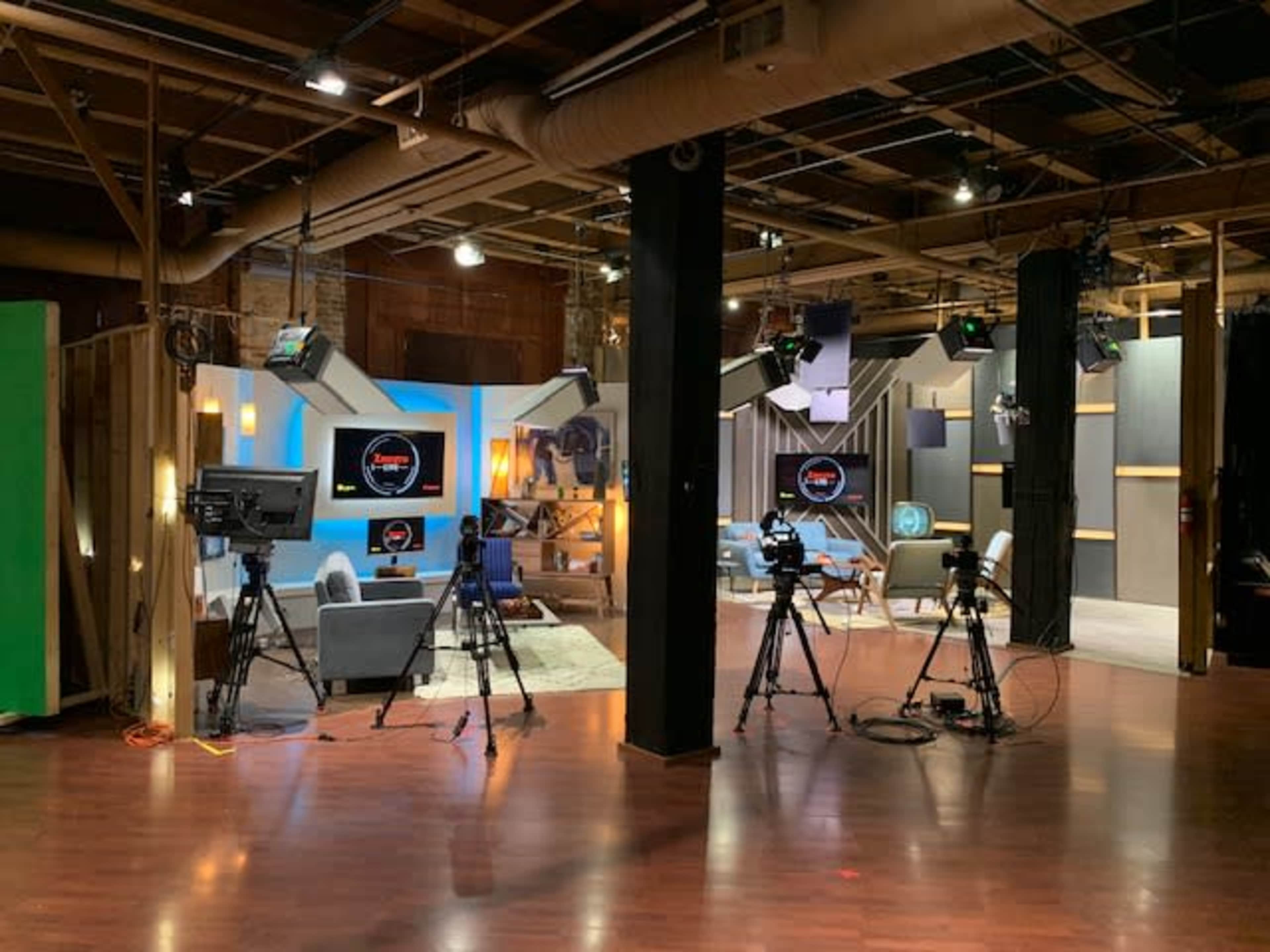 A television studio set features multiple cameras focused on a modern seating area with backdrop lighting and decorative elements.