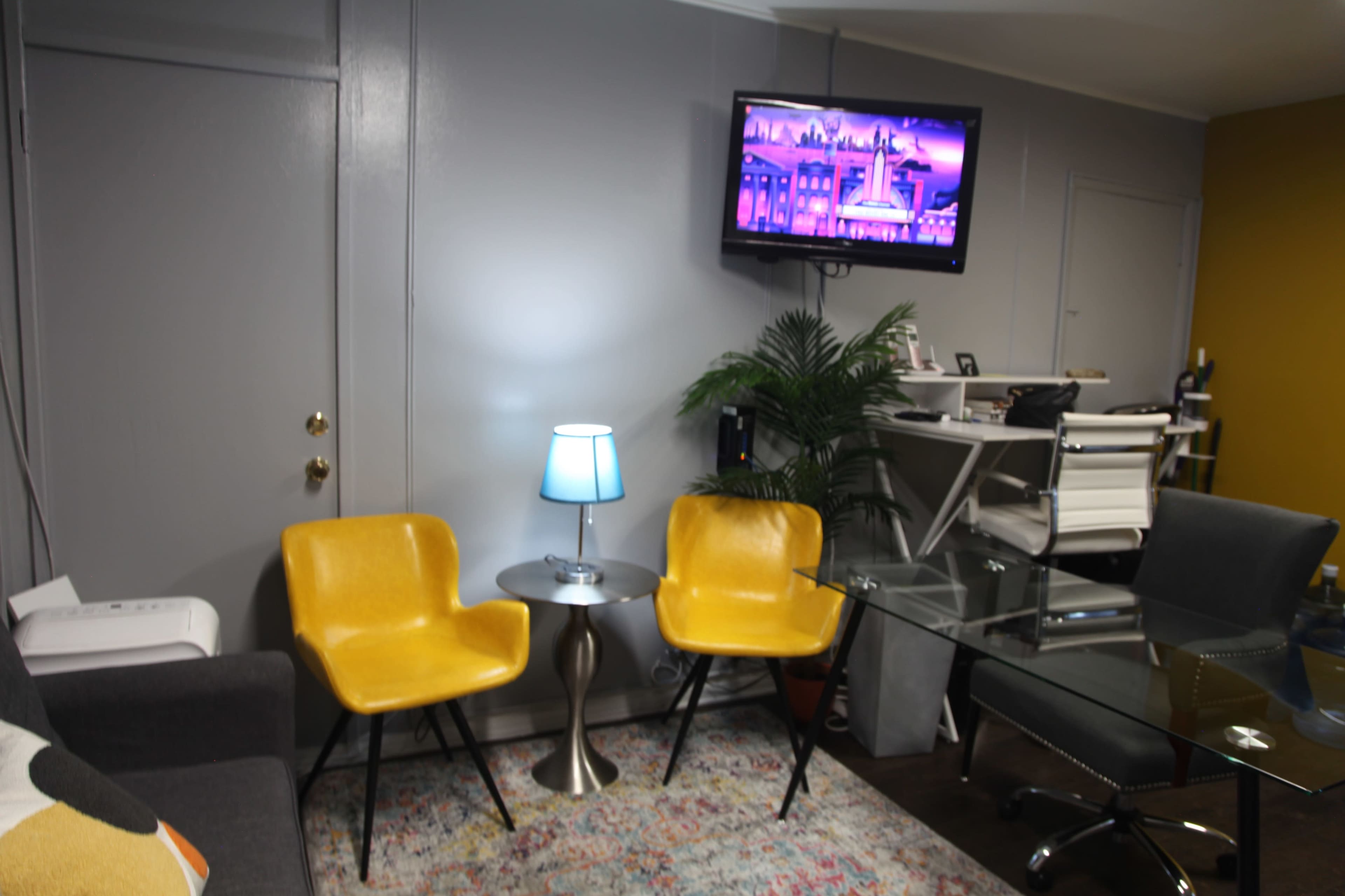 The living area features two yellow chairs, a small table with a blue lamp, a glass desk, and a television mounted on the wall.