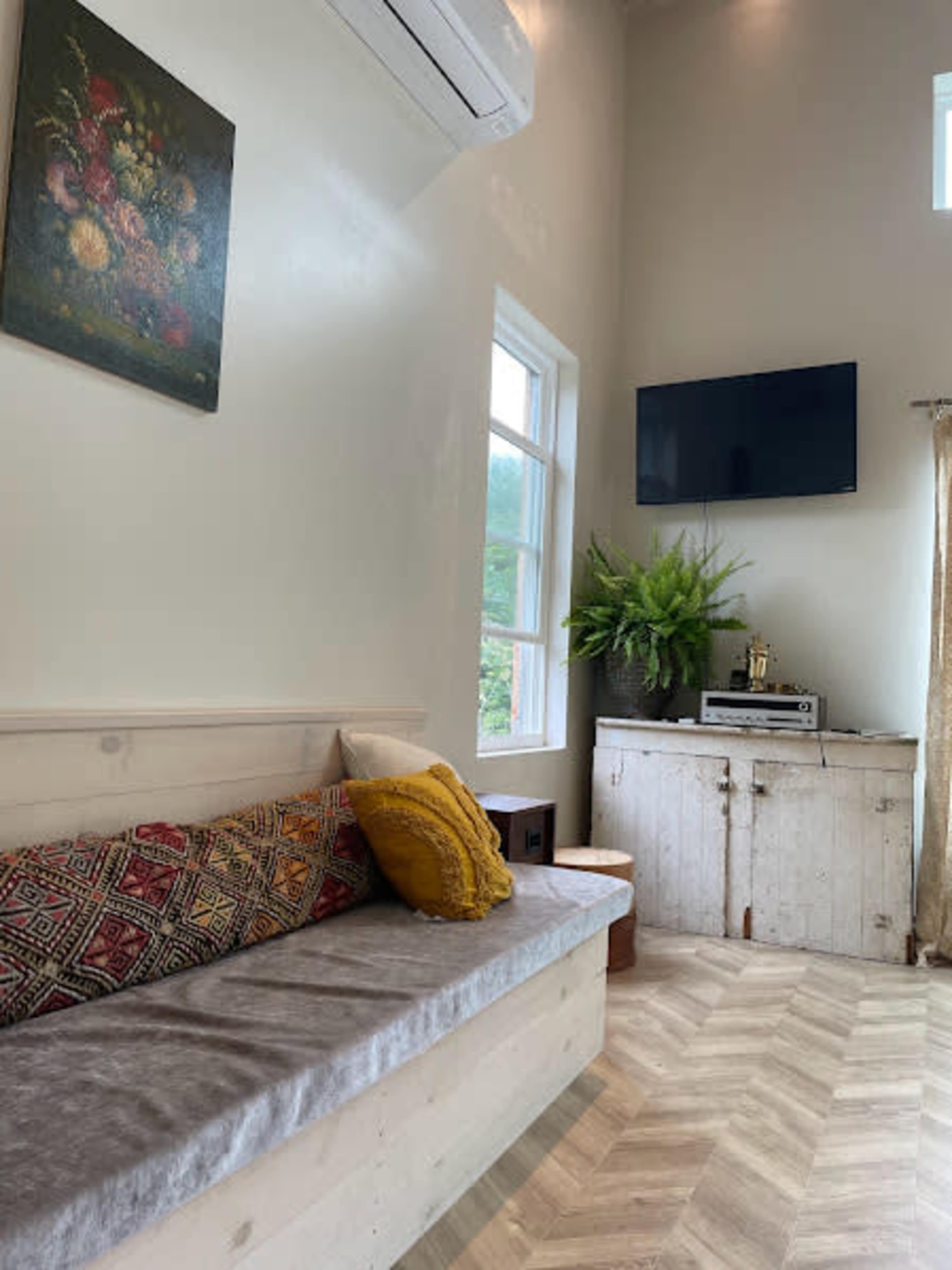 The image shows a living space with a light-colored wooden bench, decorative pillows, a television mounted on the wall, and a side cabinet with a plant beside it.