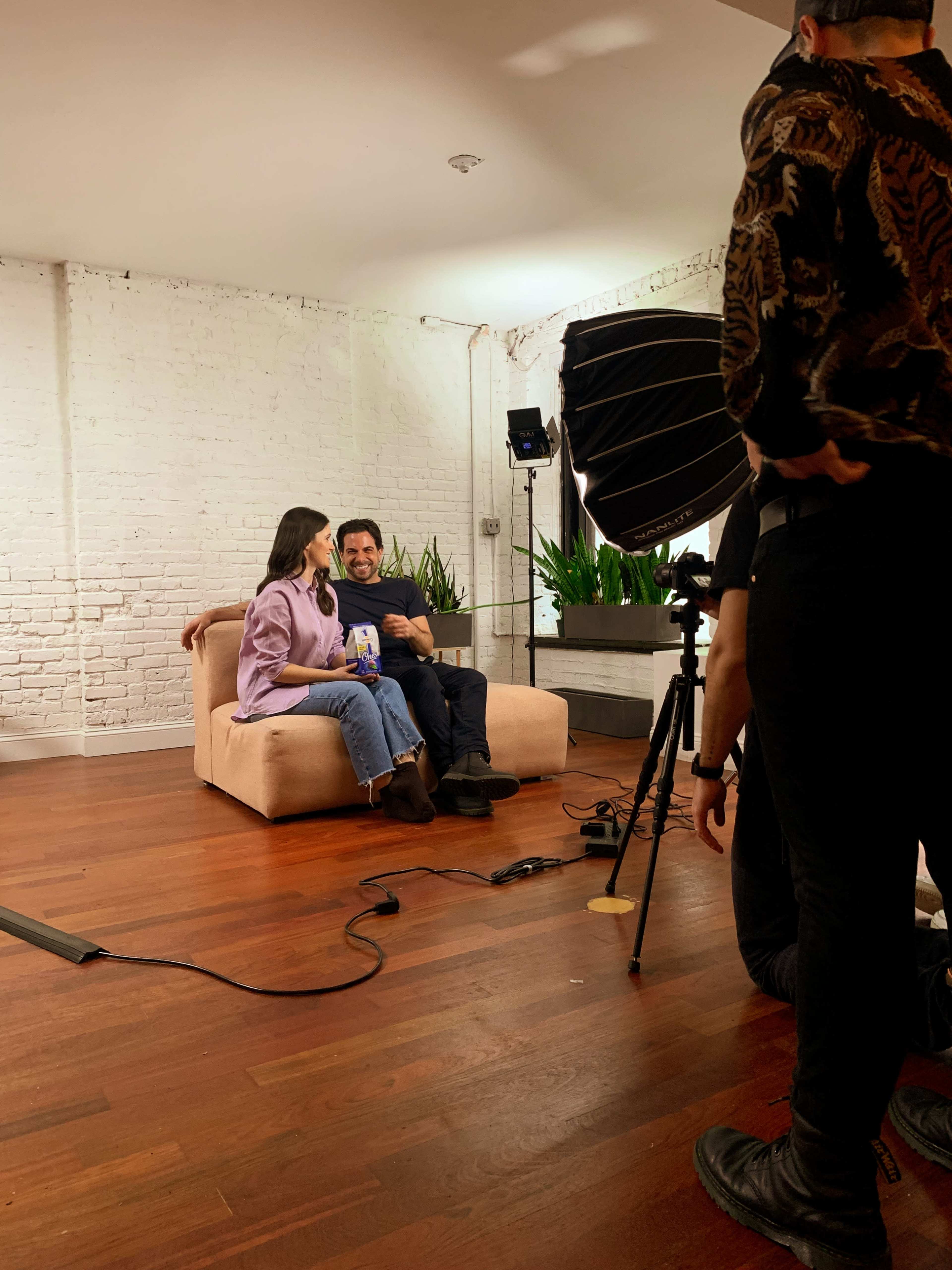 A man and a woman sit on a couch in a bright studio, engaged in conversation while a camera crew prepares to film.