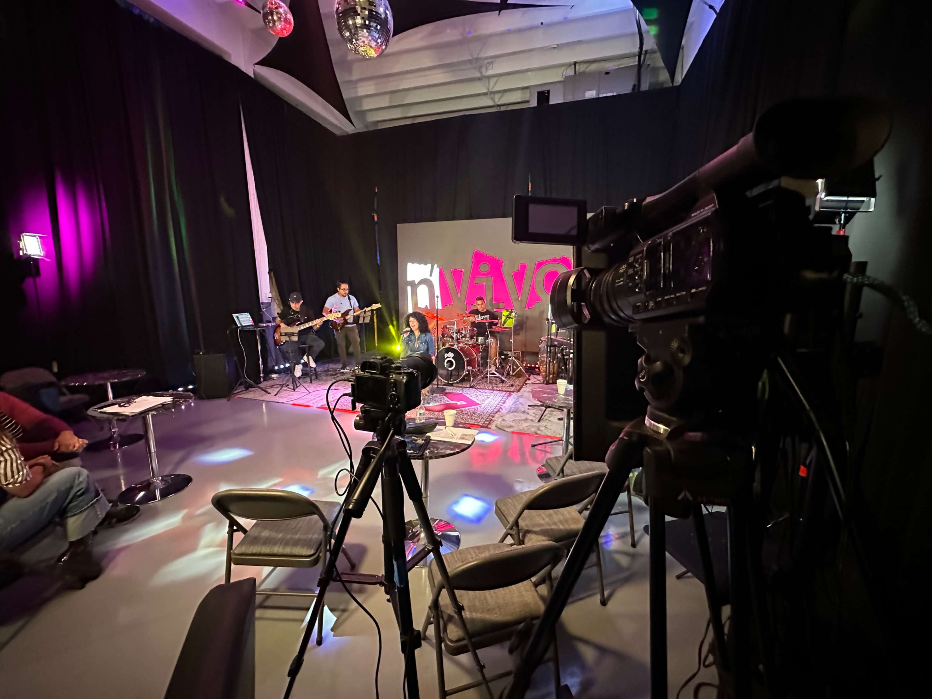 A music band performs on stage in a studio setting equipped with cameras and unique lighting.