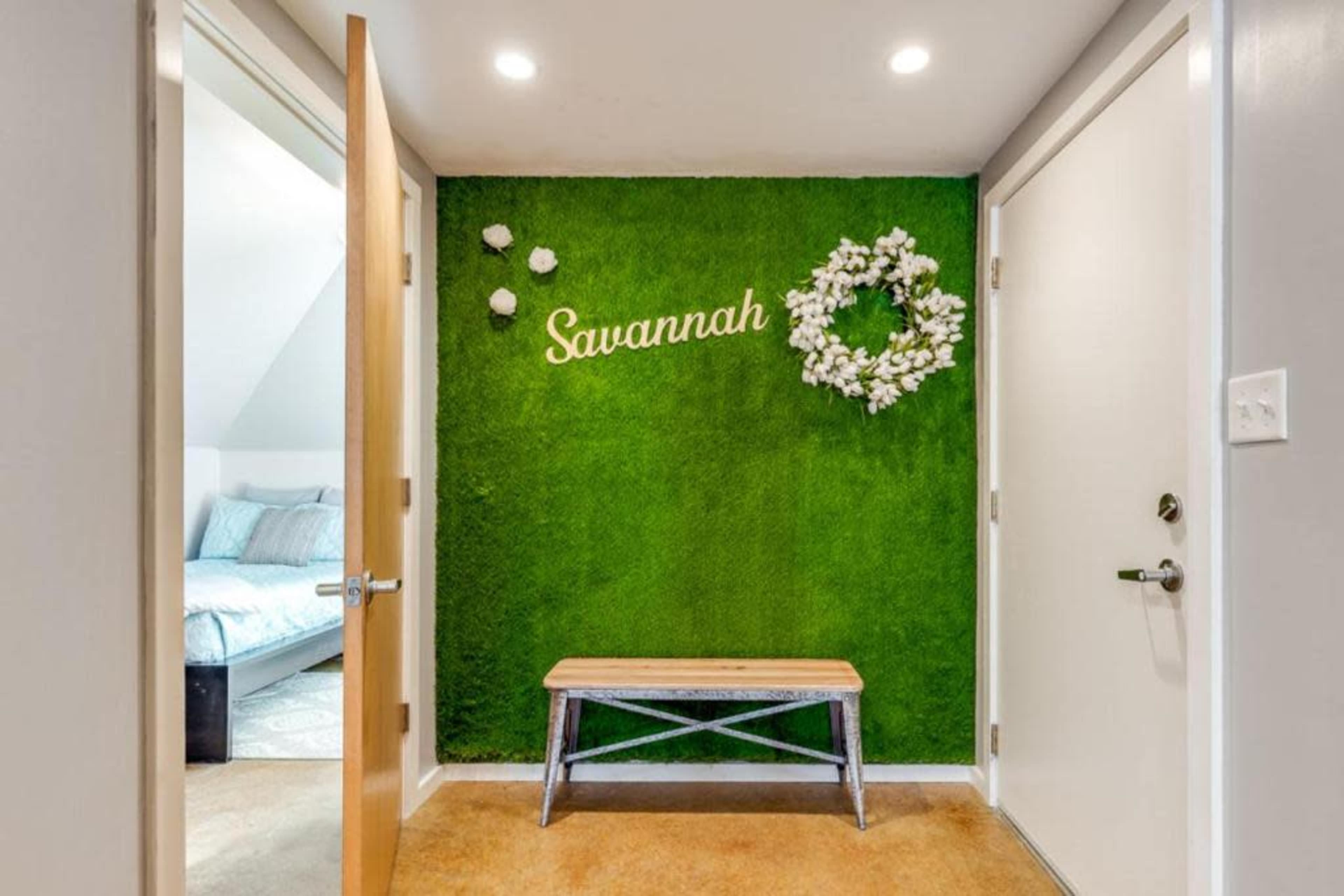 The image shows a well-lit foyer with a green moss wall featuring the name "Savannah" and a floral wreath, alongside a wooden bench and two doors.