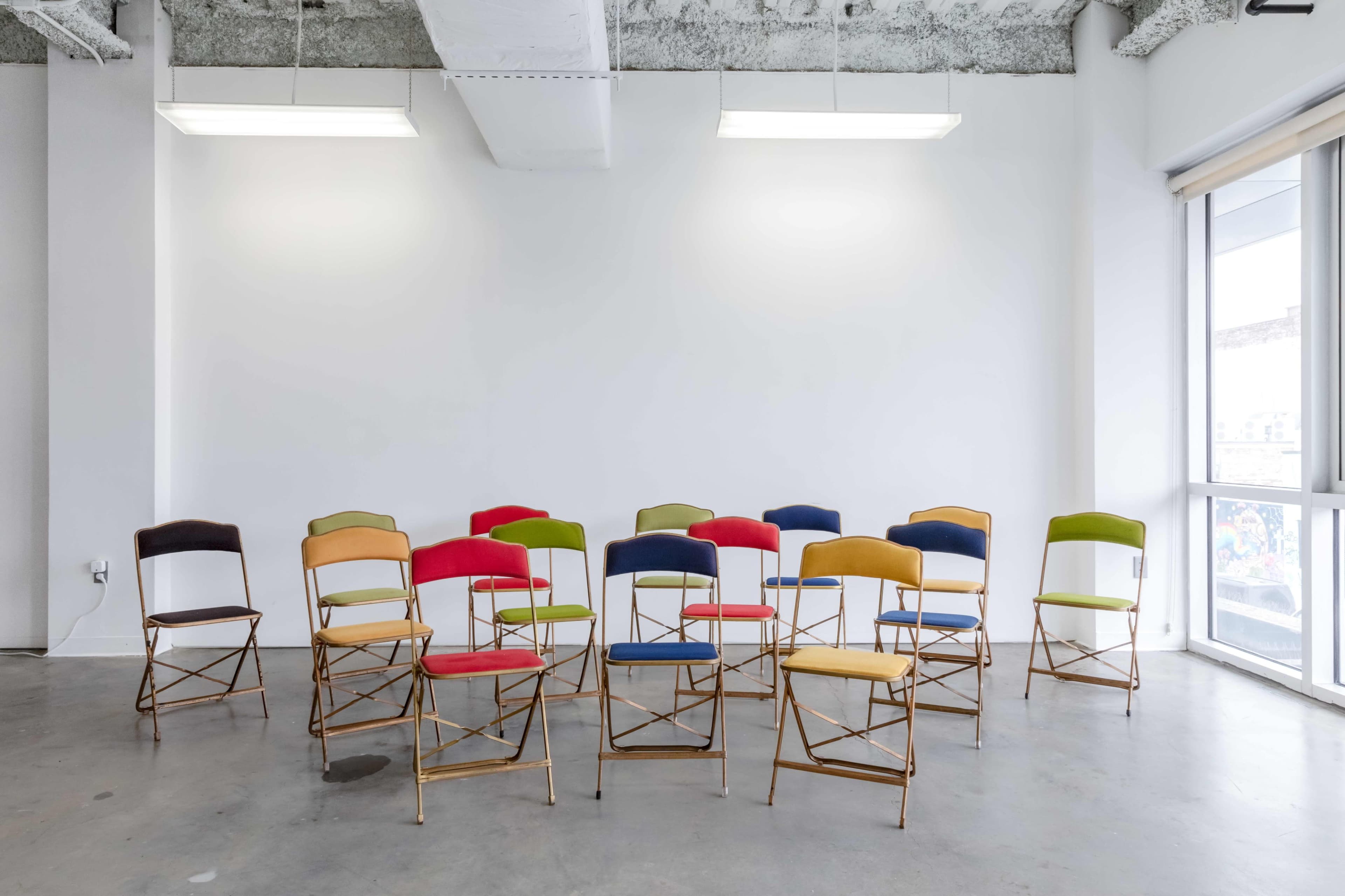 A series of colorful folding chairs are arranged in front of a white wall in a bright, minimalist room.