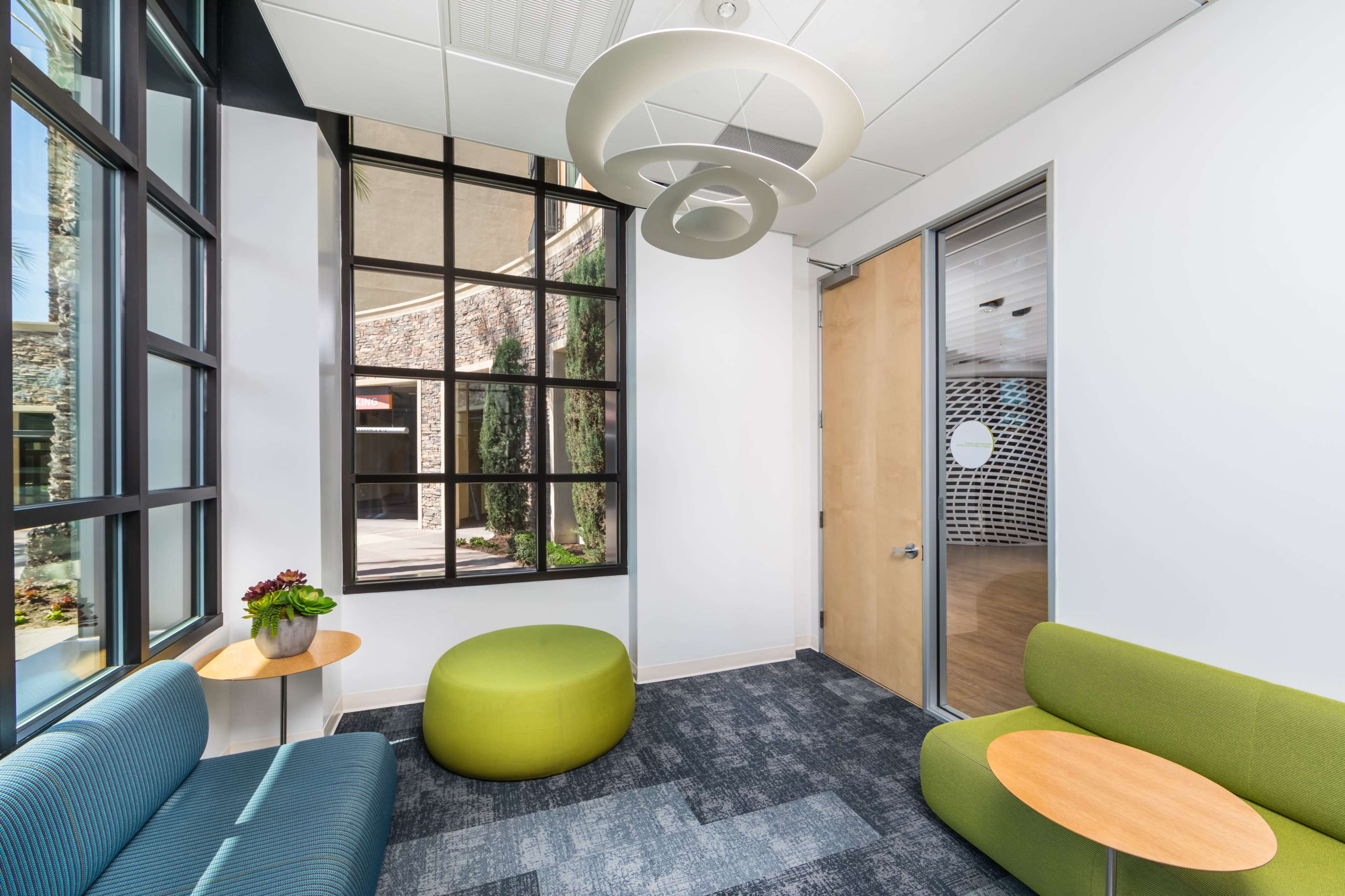 A modern lounge area with two seating options, a round green ottoman, and a small table, surrounded by large windows and a doorway leading to another room.