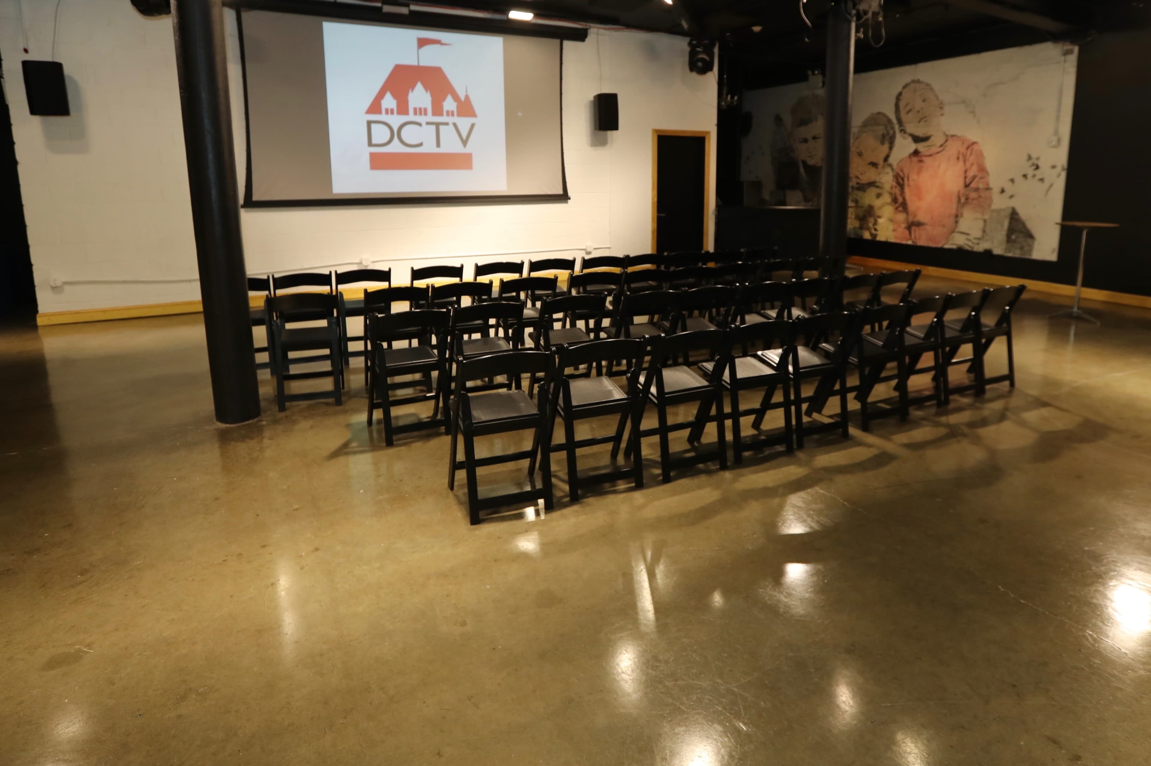 A row of black folding chairs is arranged in front of a large screen displaying the DCTV logo in a polished concrete room.