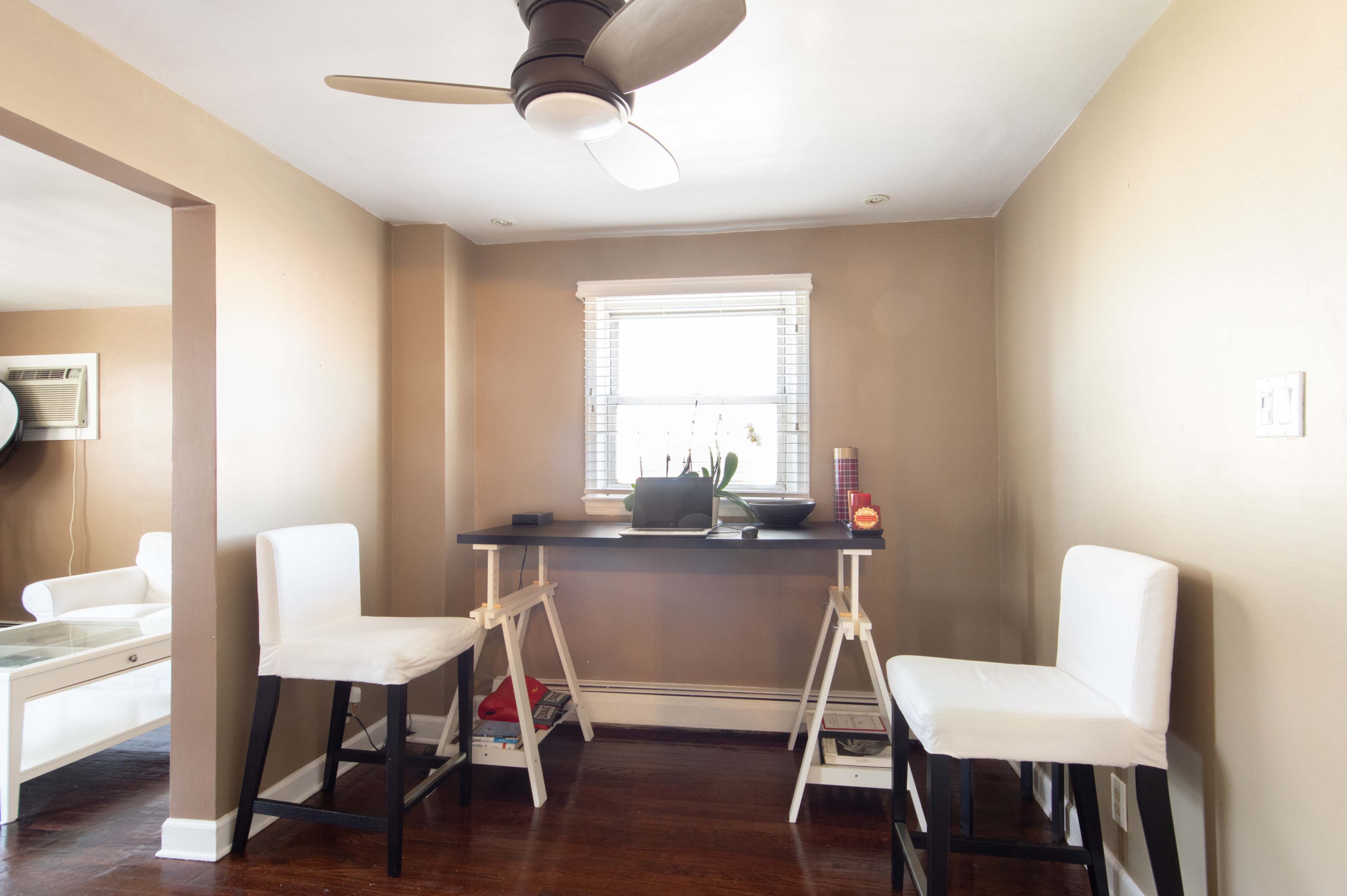 The image shows a small room with two white barstools positioned at a narrow table beneath a window with a plant on it.