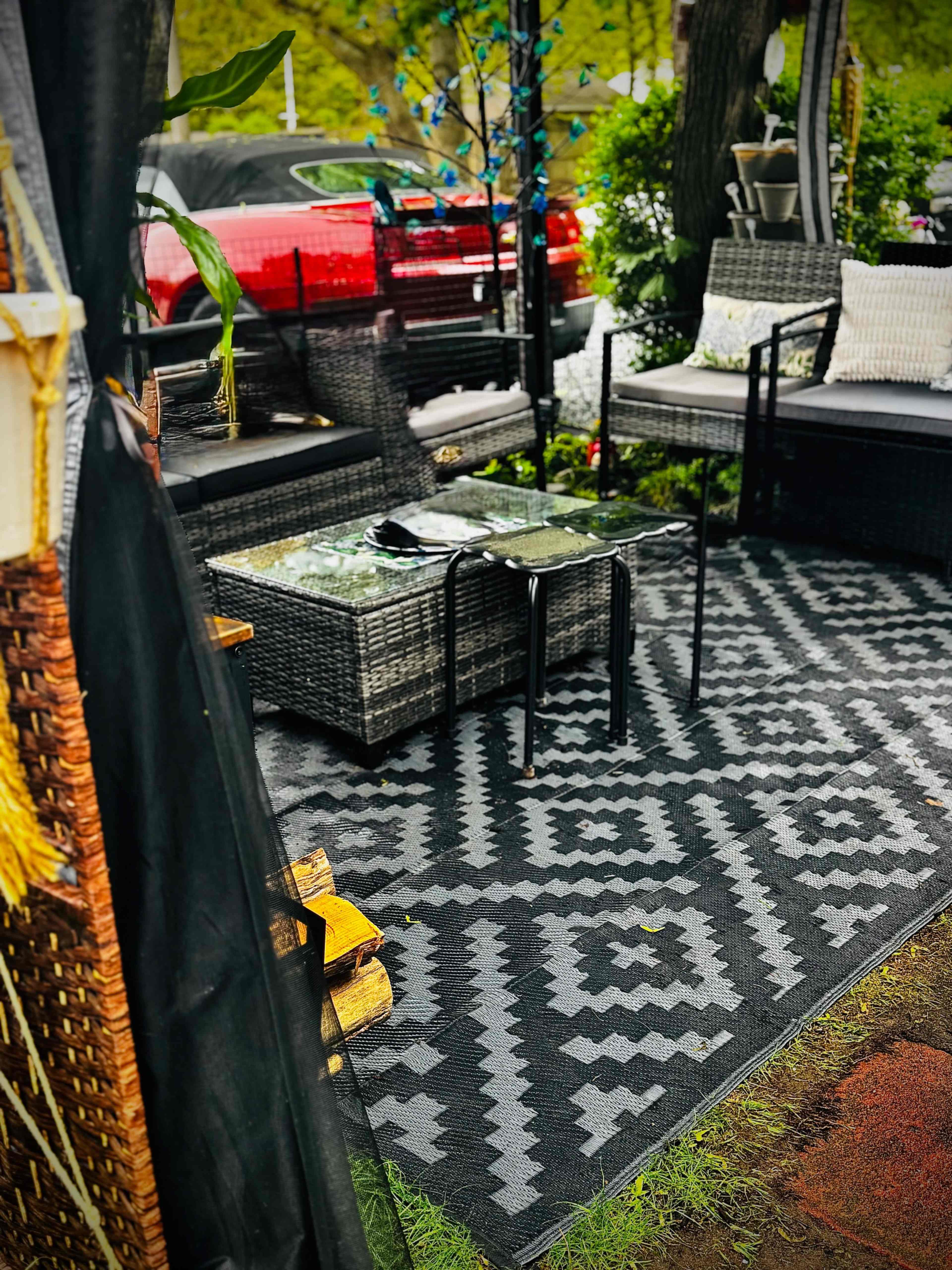 A cozy outdoor seating area features wicker furniture arranged around a patterned rug, with a car visible in the background.