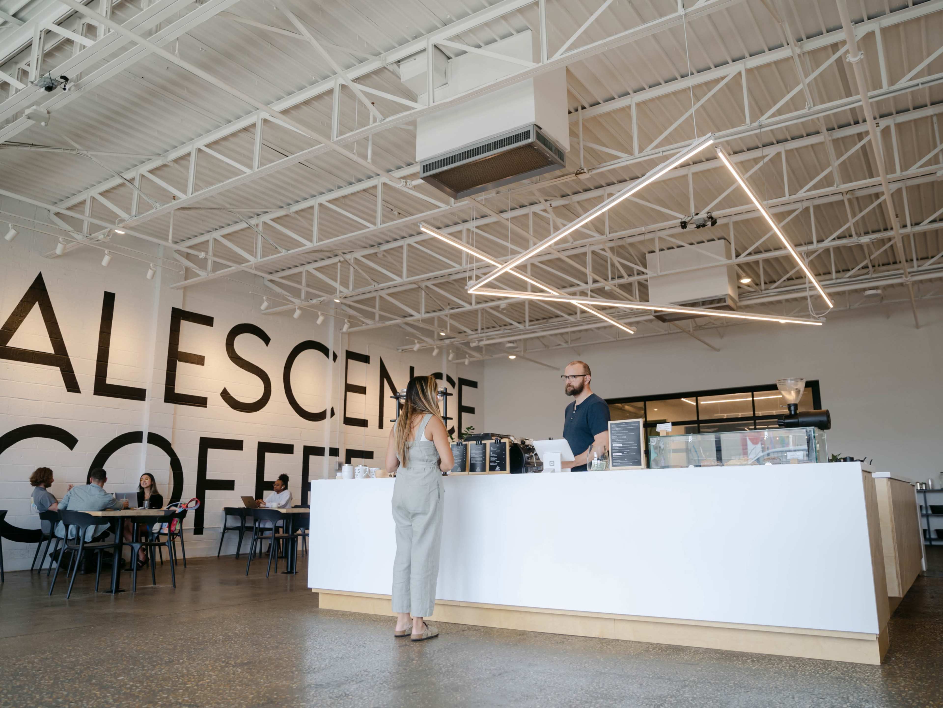 A coffee shop features a large white counter with a barista serving a customer, while several patrons sit at tables in the background.