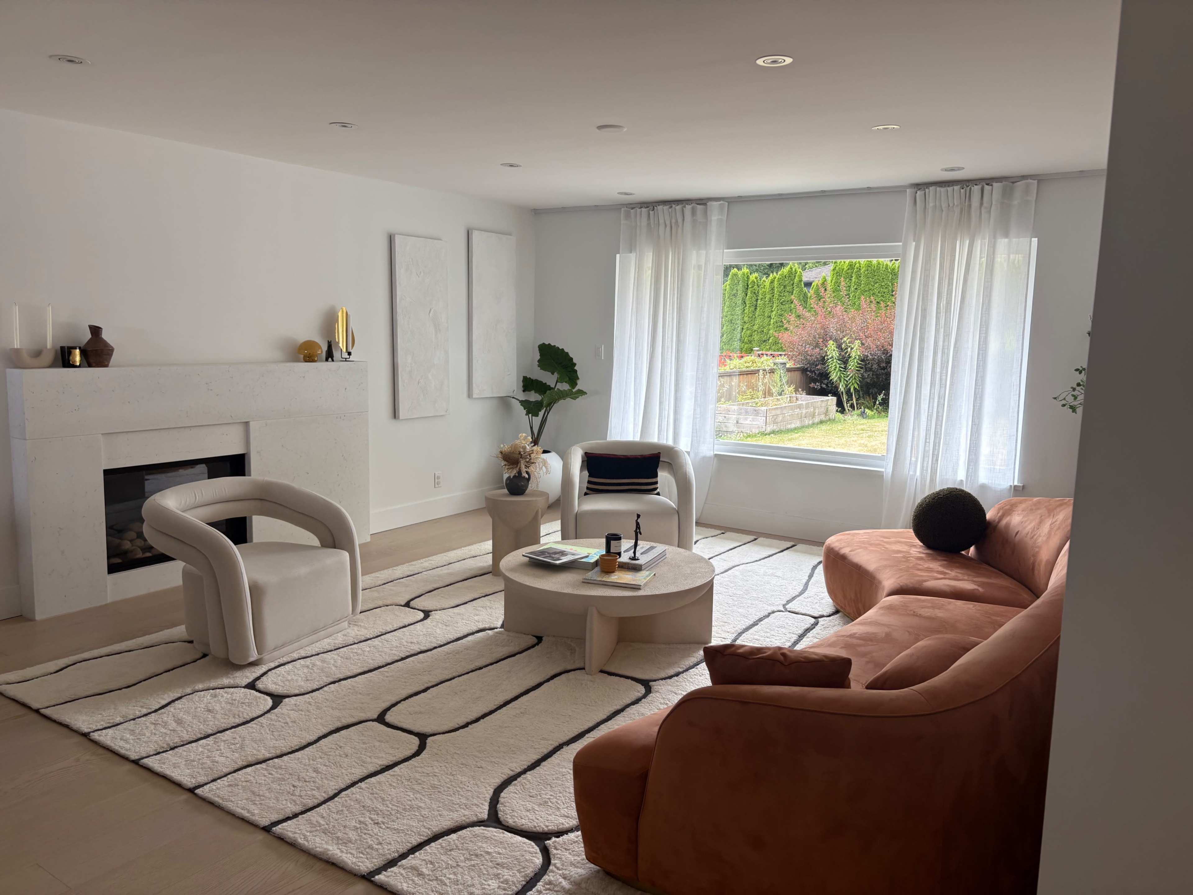 The living room features a modern design with a curved orange sofa, white armchairs, a circular coffee table, and large windows letting in natural light.