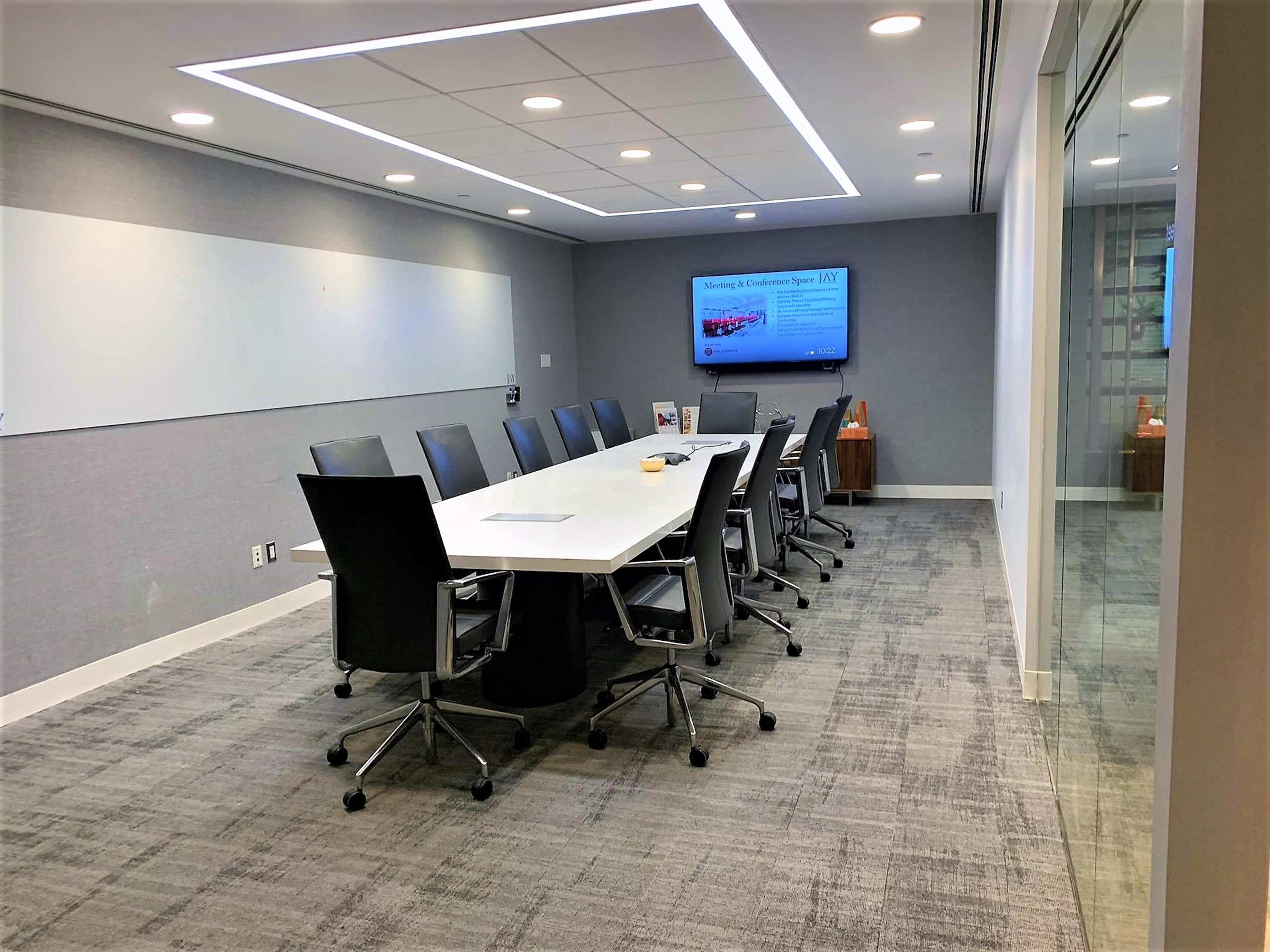 A modern conference room features a large white table surrounded by black chairs, with a presentation screen on the wall and a glass partition.