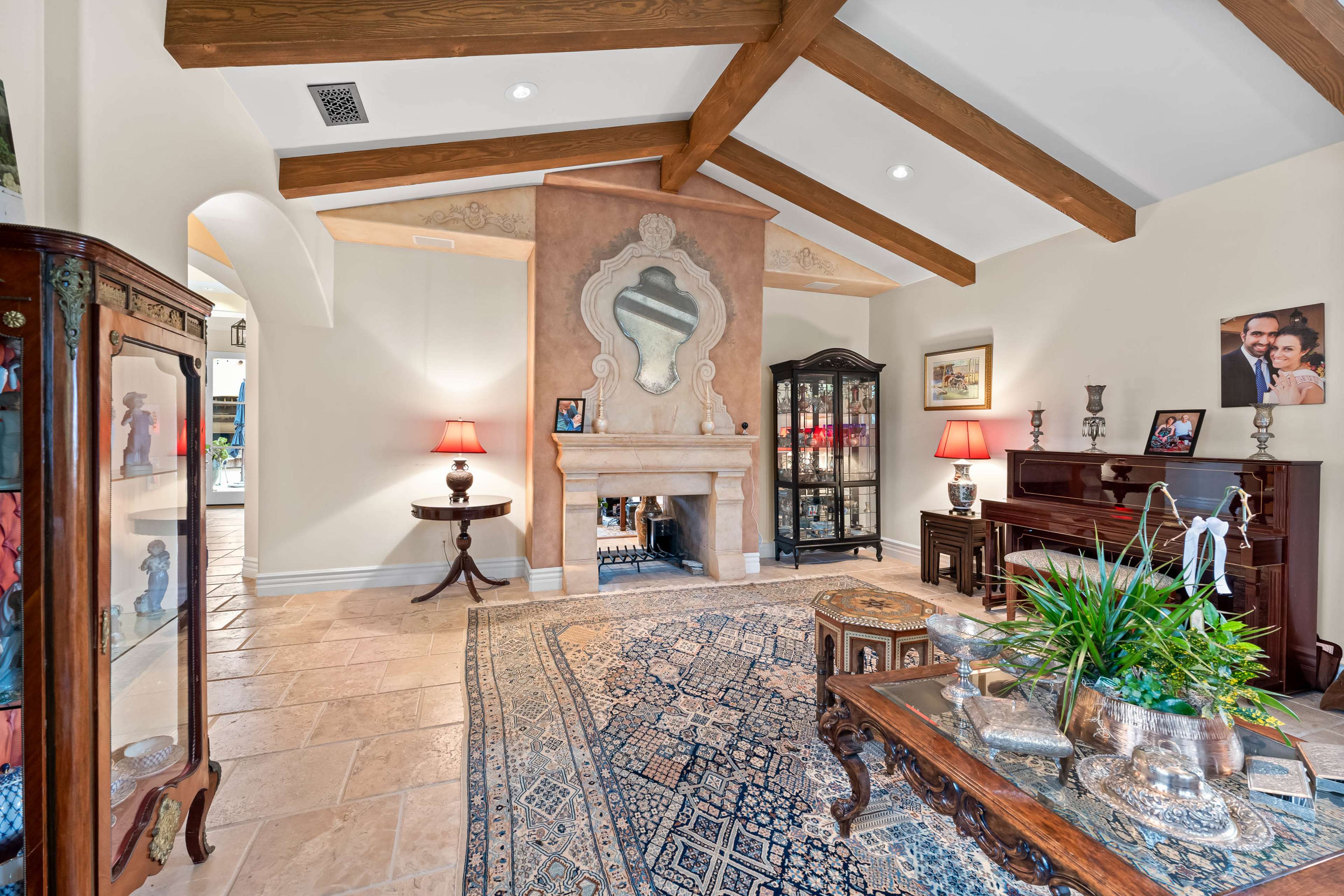 The living room features a fireplace with a decorative mantle, wooden beams on the ceiling, and various furnishings including a piano and display cabinets.