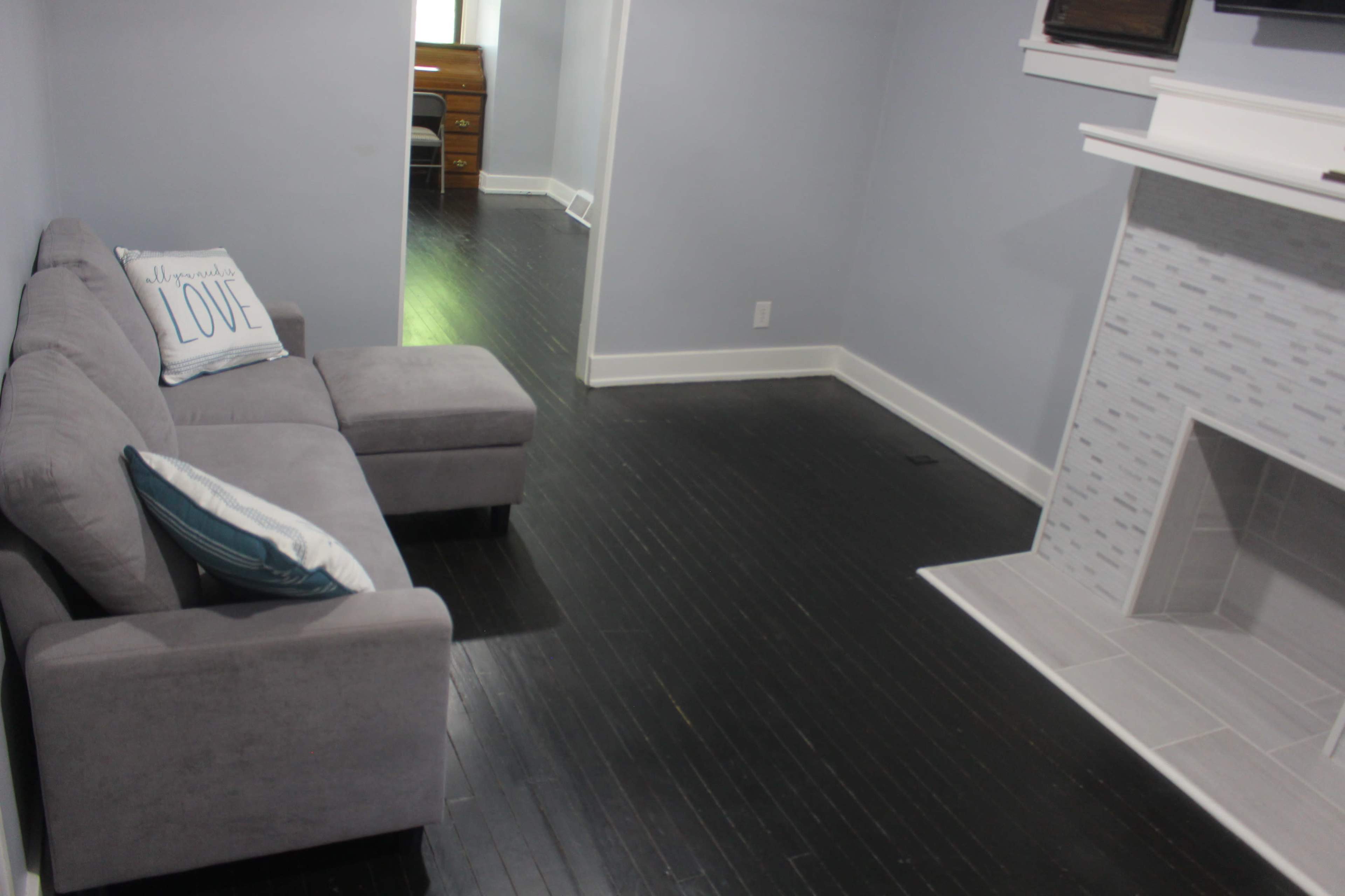 A gray sectional sofa sits on a dark hardwood floor in a light-colored room with a white fireplace and a doorway leading to another area.