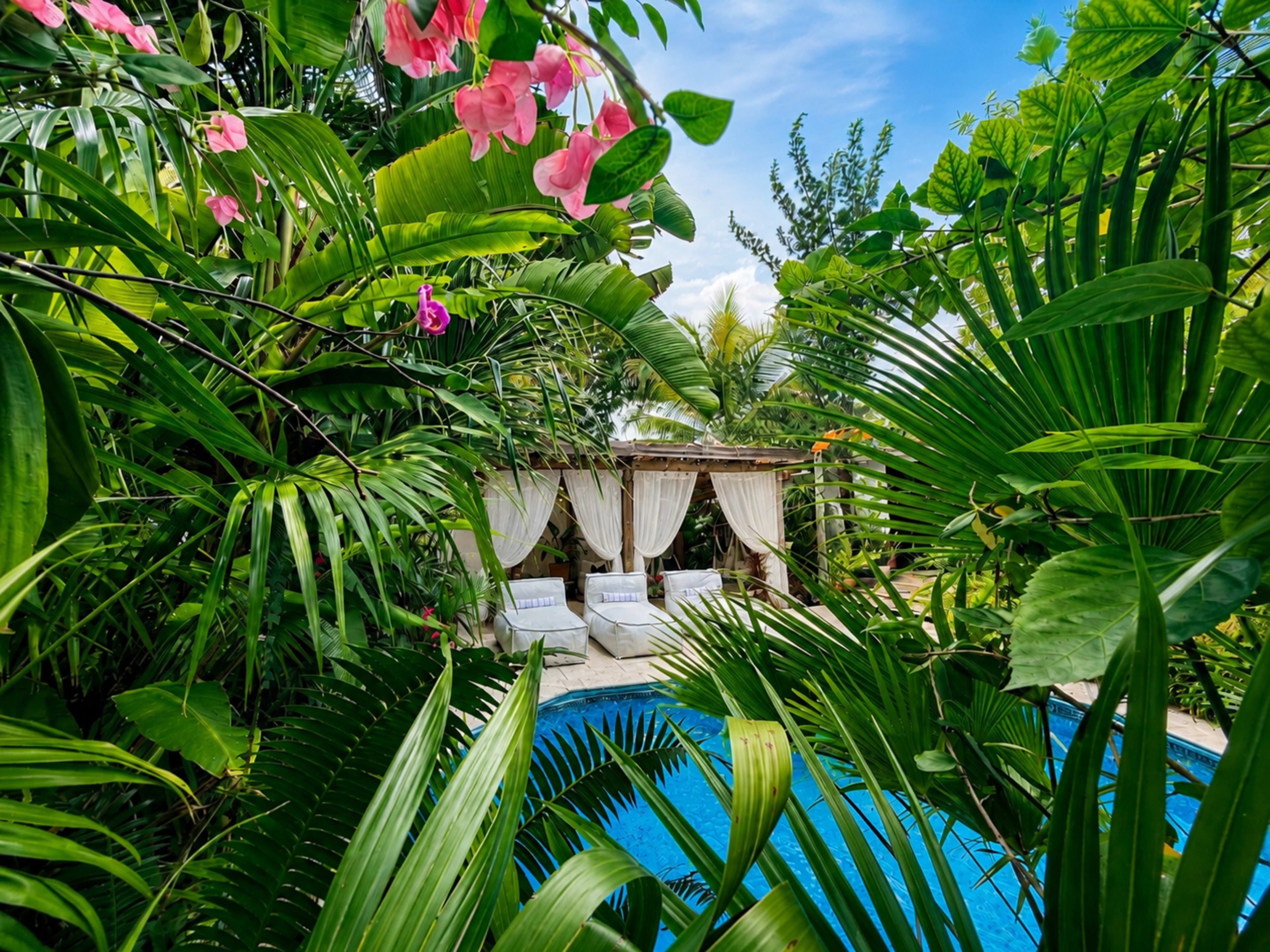 A serene pool area is surrounded by lush tropical plants and flowers, featuring two sun loungers under a canopy.