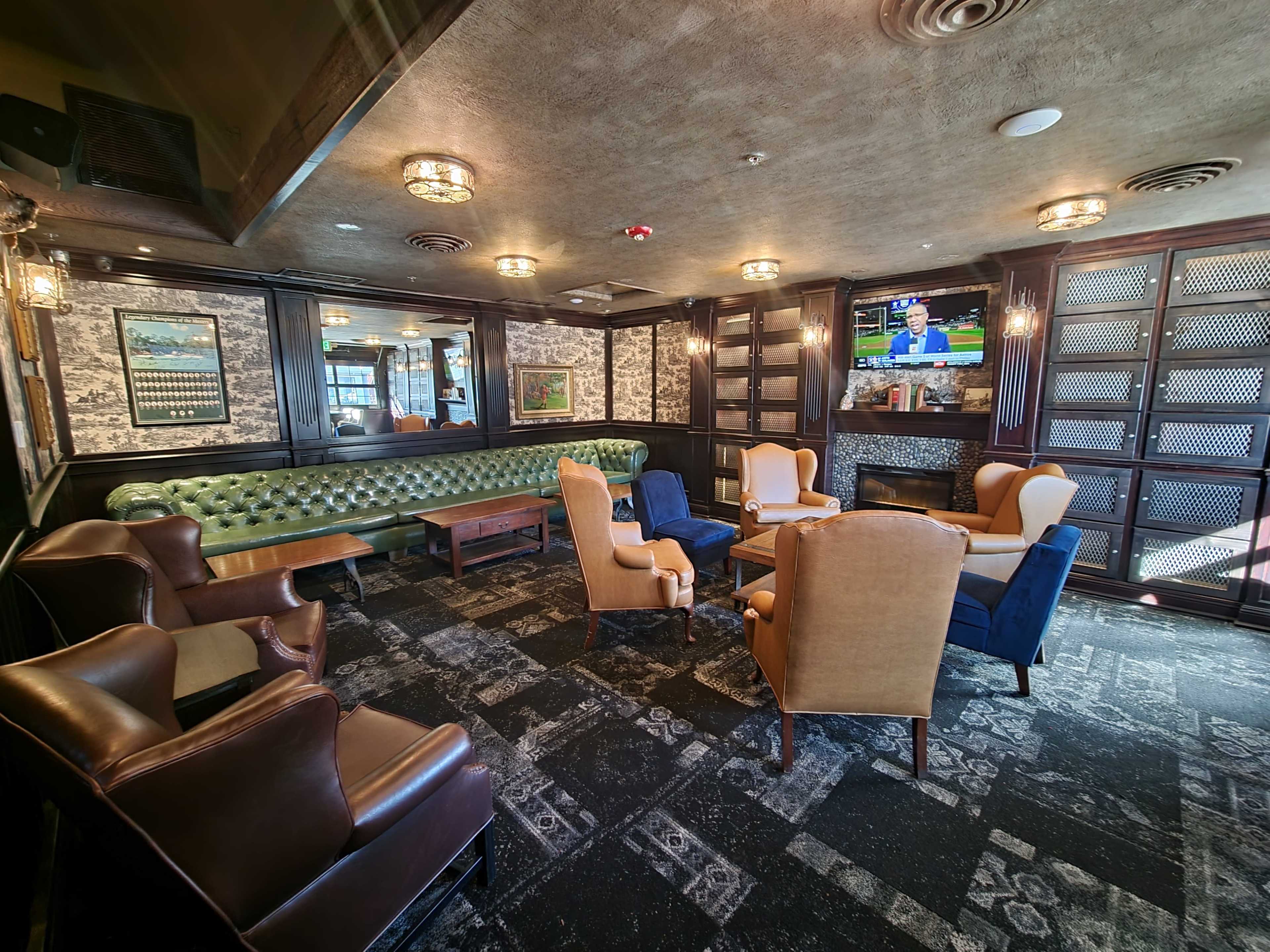 A cozy lounge area features a green tufted sofa, several armchairs, and a television mounted on the wall.