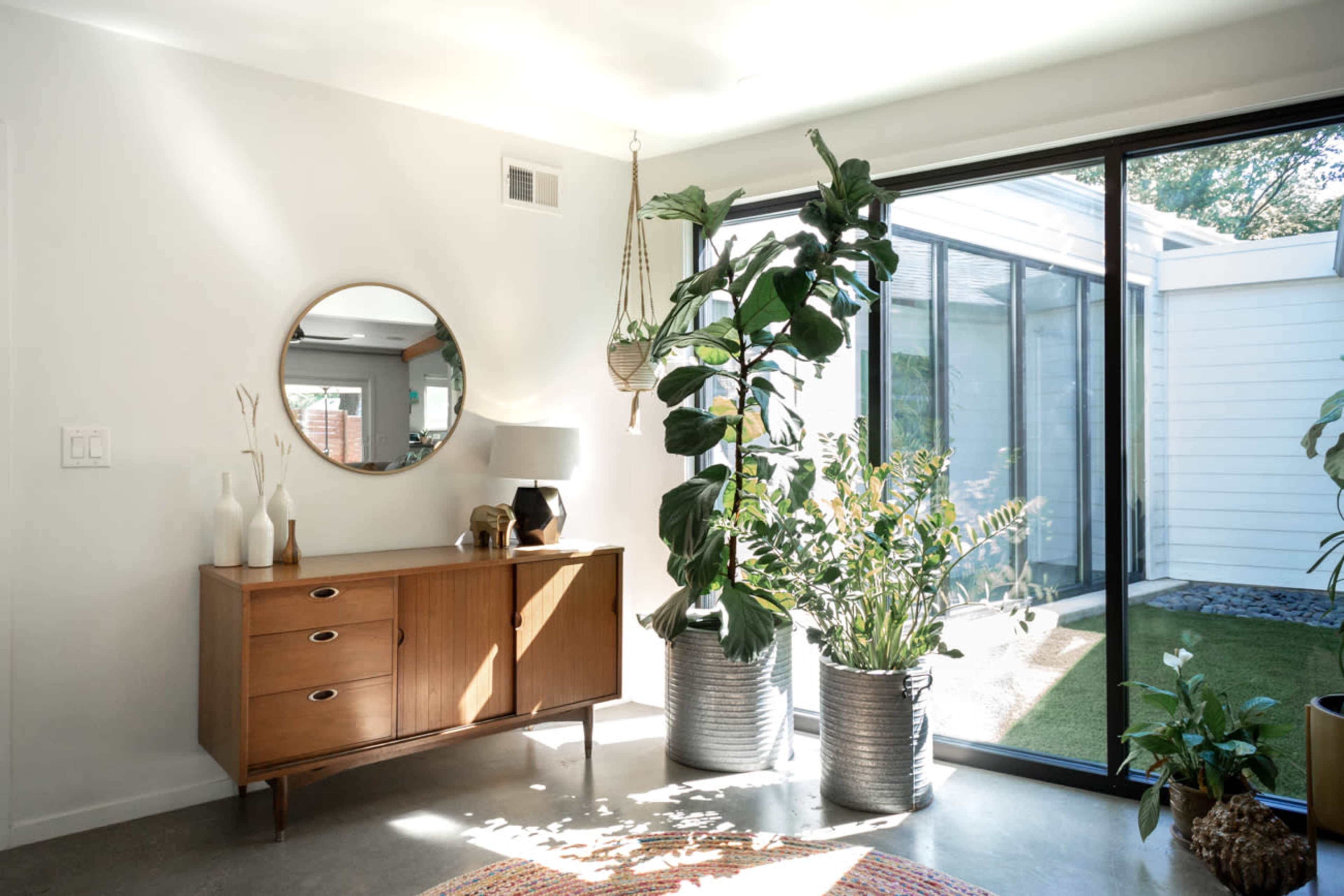 A bright entryway features a wooden sideboard, a round mirror, large indoor plants, and a glass wall that opens to a small garden.