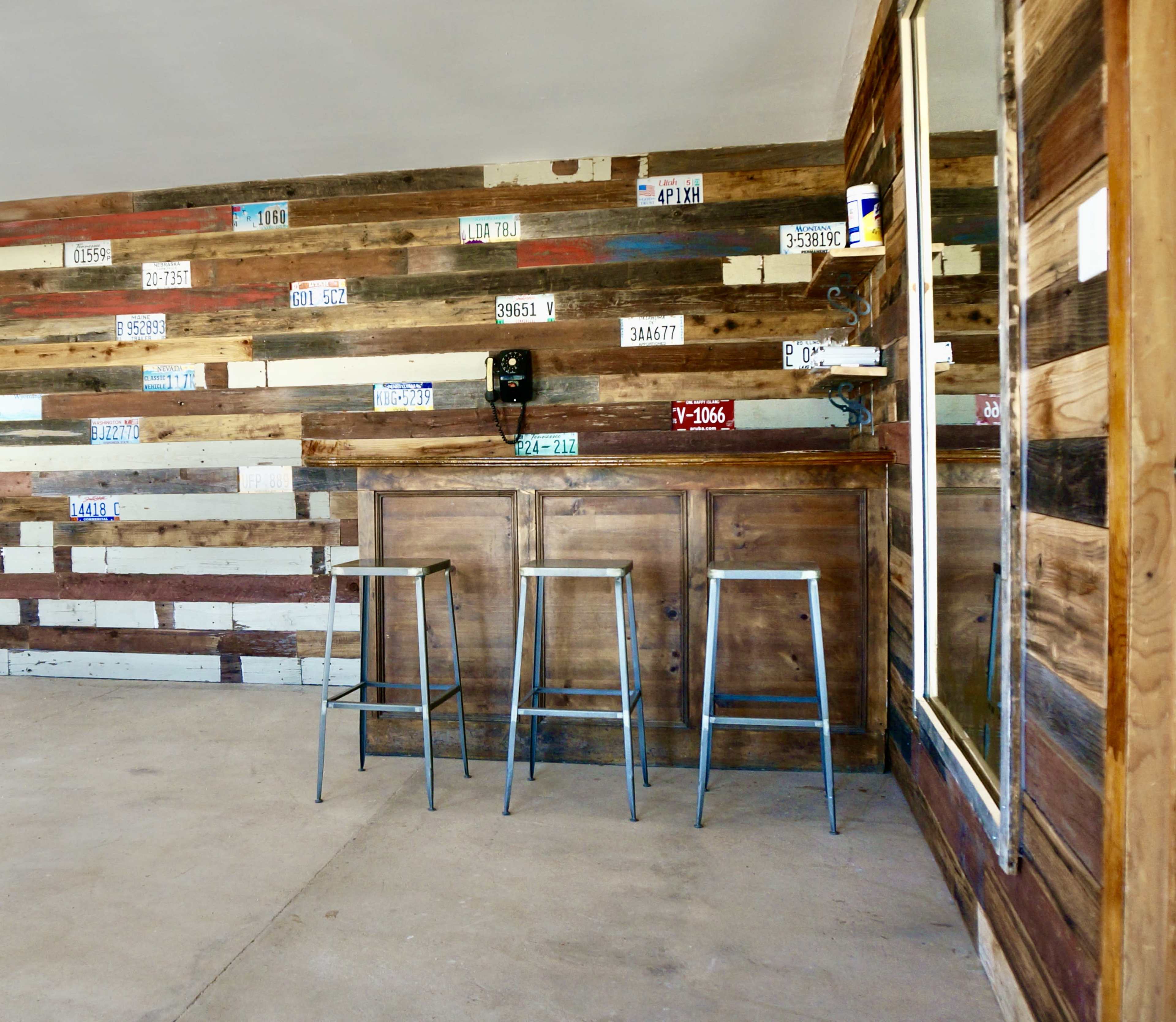 The image shows a rustic interior with a wooden bar, three metal stools, and a wall decorated with various license plates.