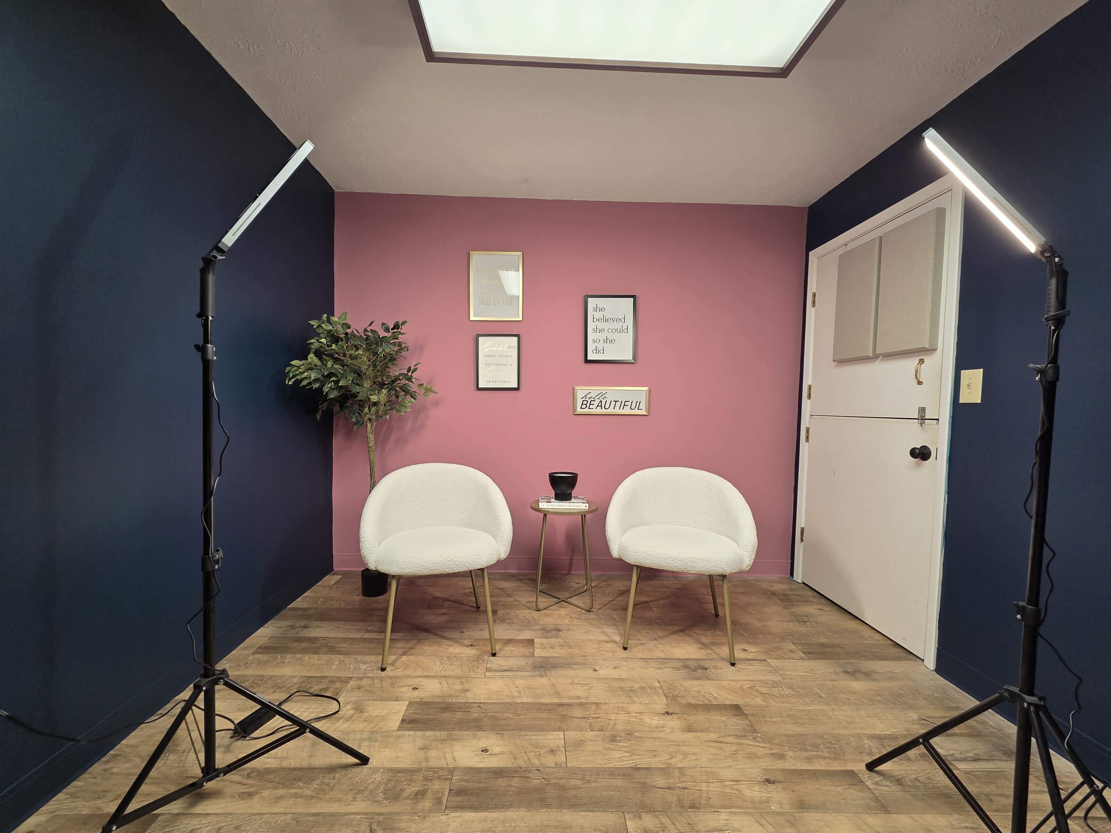 The image shows a small, staged room with two white chairs, a round table, and decorative wall art, illuminated by lights on stands.