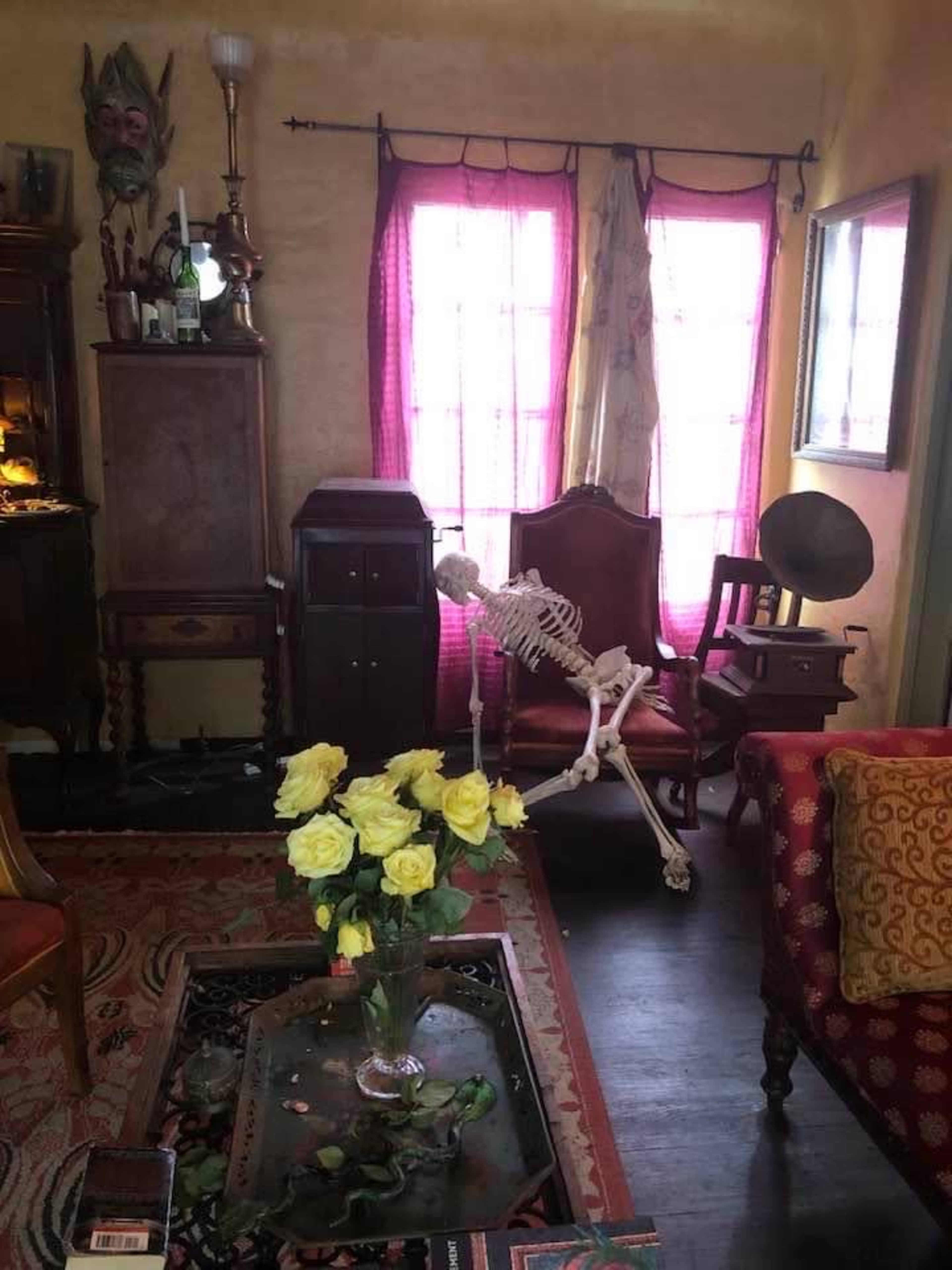 A skeleton sits in an ornate chair next to a vase of yellow roses in a vintage-style room with wooden furniture and colorful curtains.