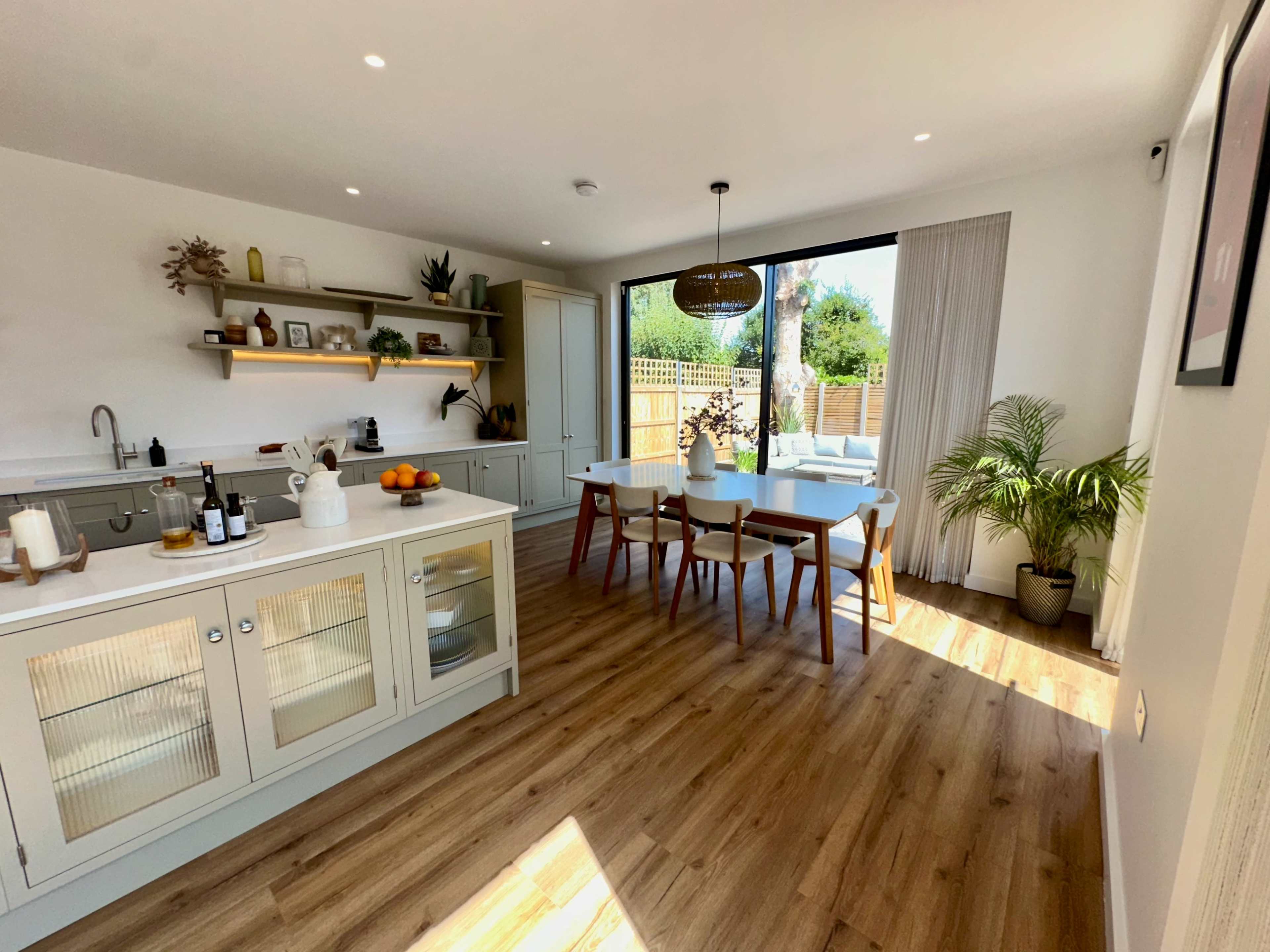 The image shows a modern kitchen and dining area with a large window allowing natural light to illuminate the space, featuring a dining table and chairs, and a kitchen island with cabinets.